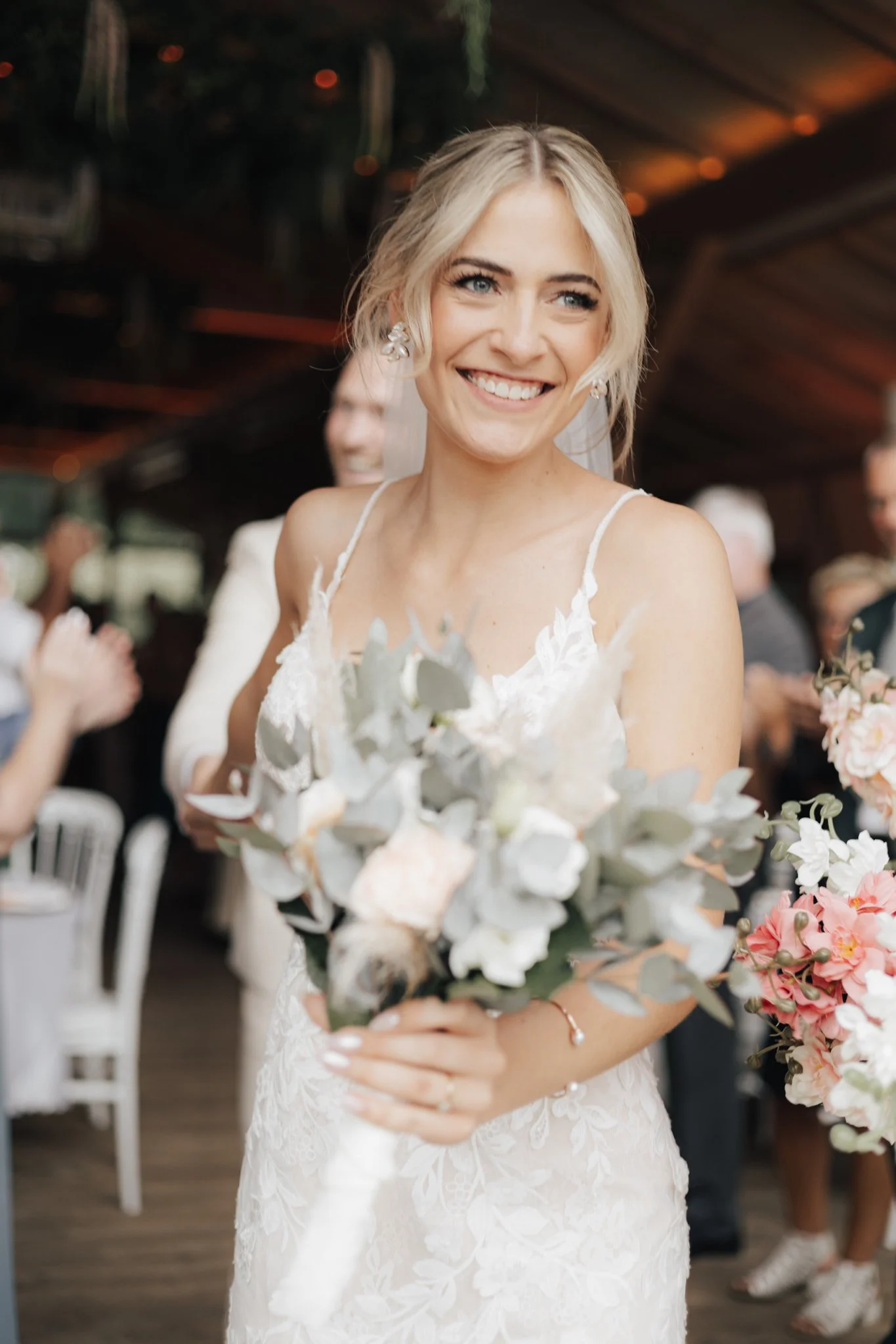 Eine lächelnde Braut in einem weißen Hochzeitskleid hält einen Blumenstrauß mit weißen und grünen Pflanzen, im Hintergrund sitzen Gäste an einem festlich gedeckten Tisch.