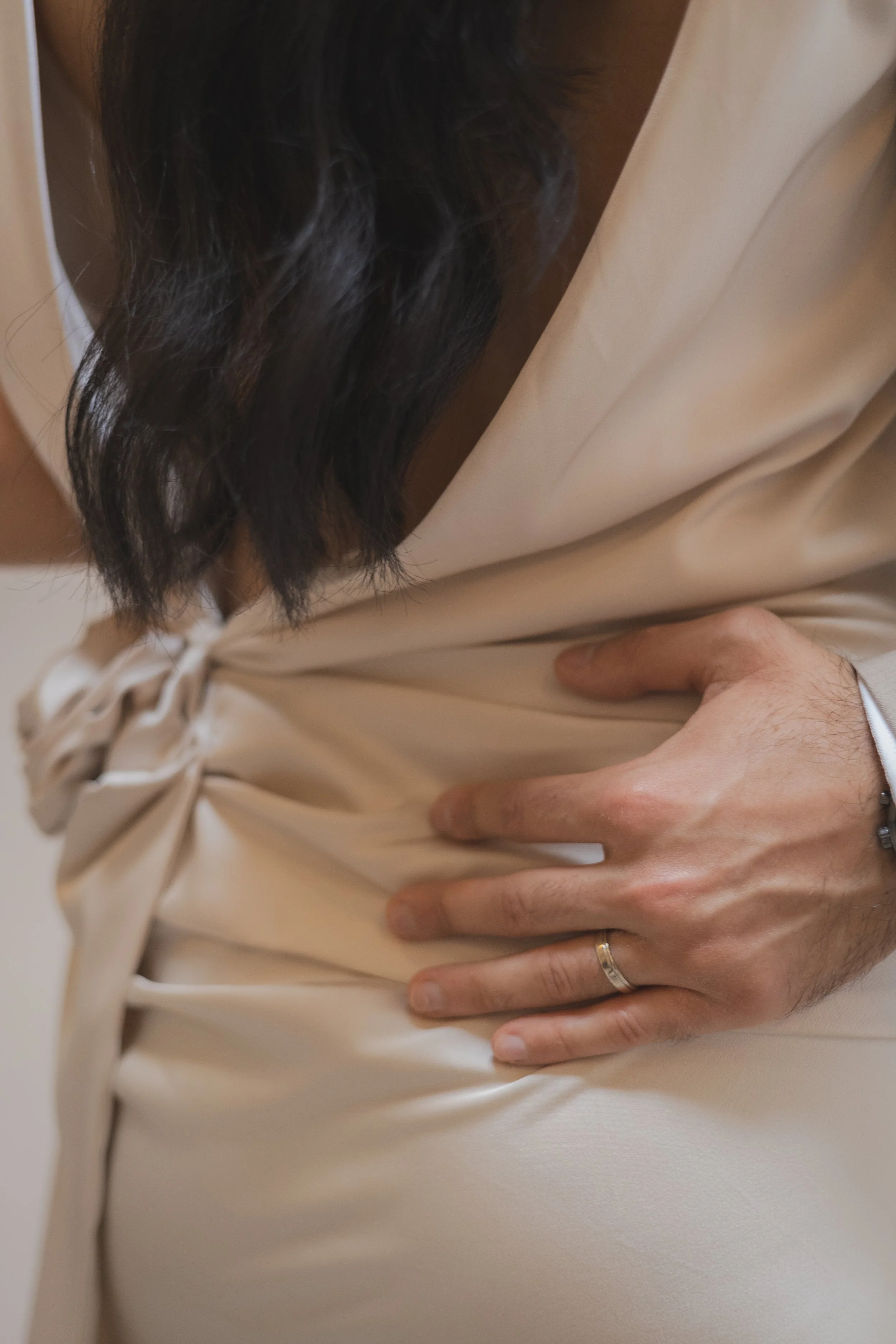 Ein Mensch mit dunklen Haaren trägt ein beiges Kleid und hält seine Hand auf den Bauch. Der Fokus liegt auf der Hand und dem Bauchbereich.