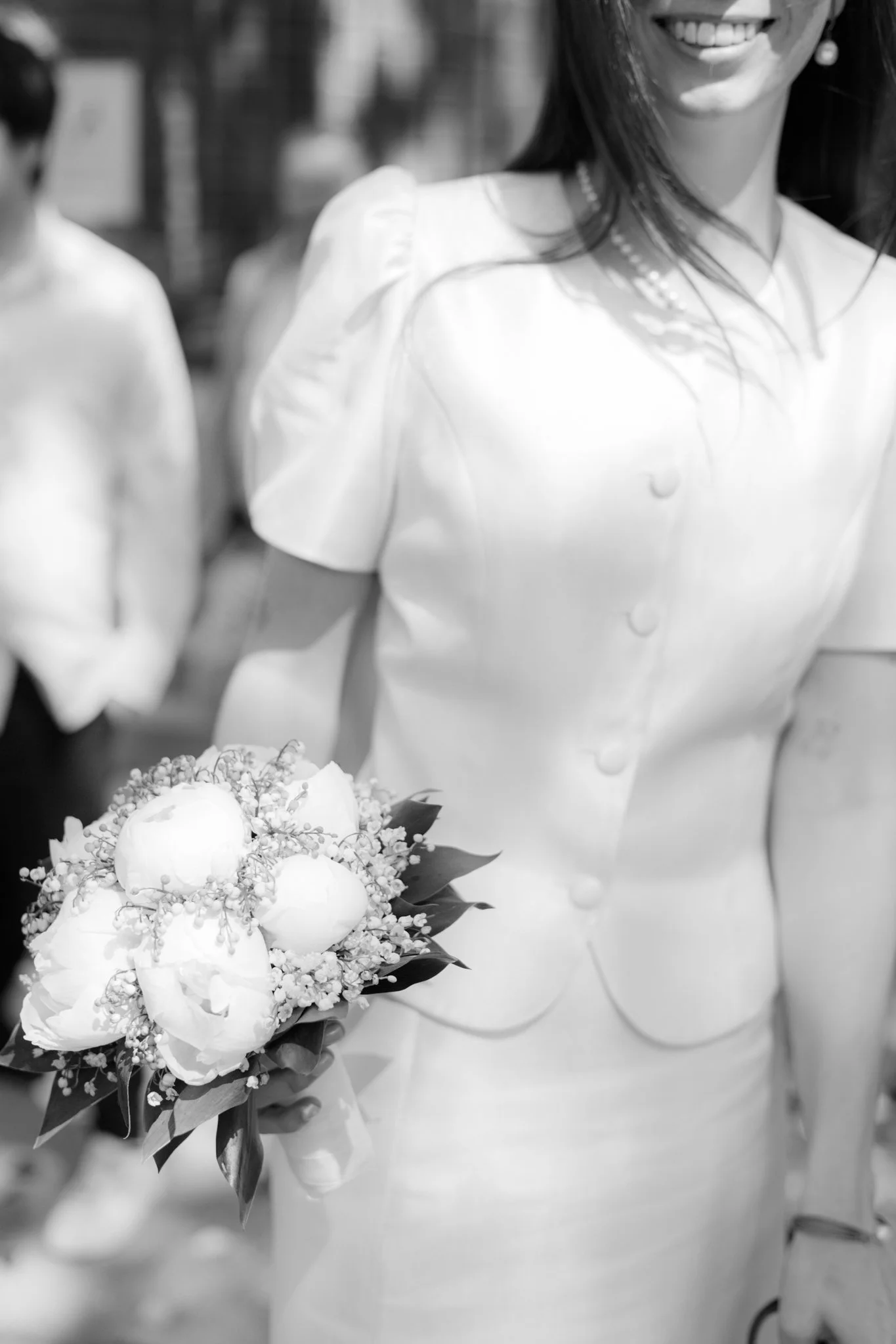 Schwarz-weiß-Foto einer Hochzeits- oder Festtagsbraut, die ein Brautstrauß hält, und sie trägt ein weißes Kleid mit Knöpfen auf der Vorderseite. Sie lächelt und trägt Perlenohrringe und eine Perlenkette.