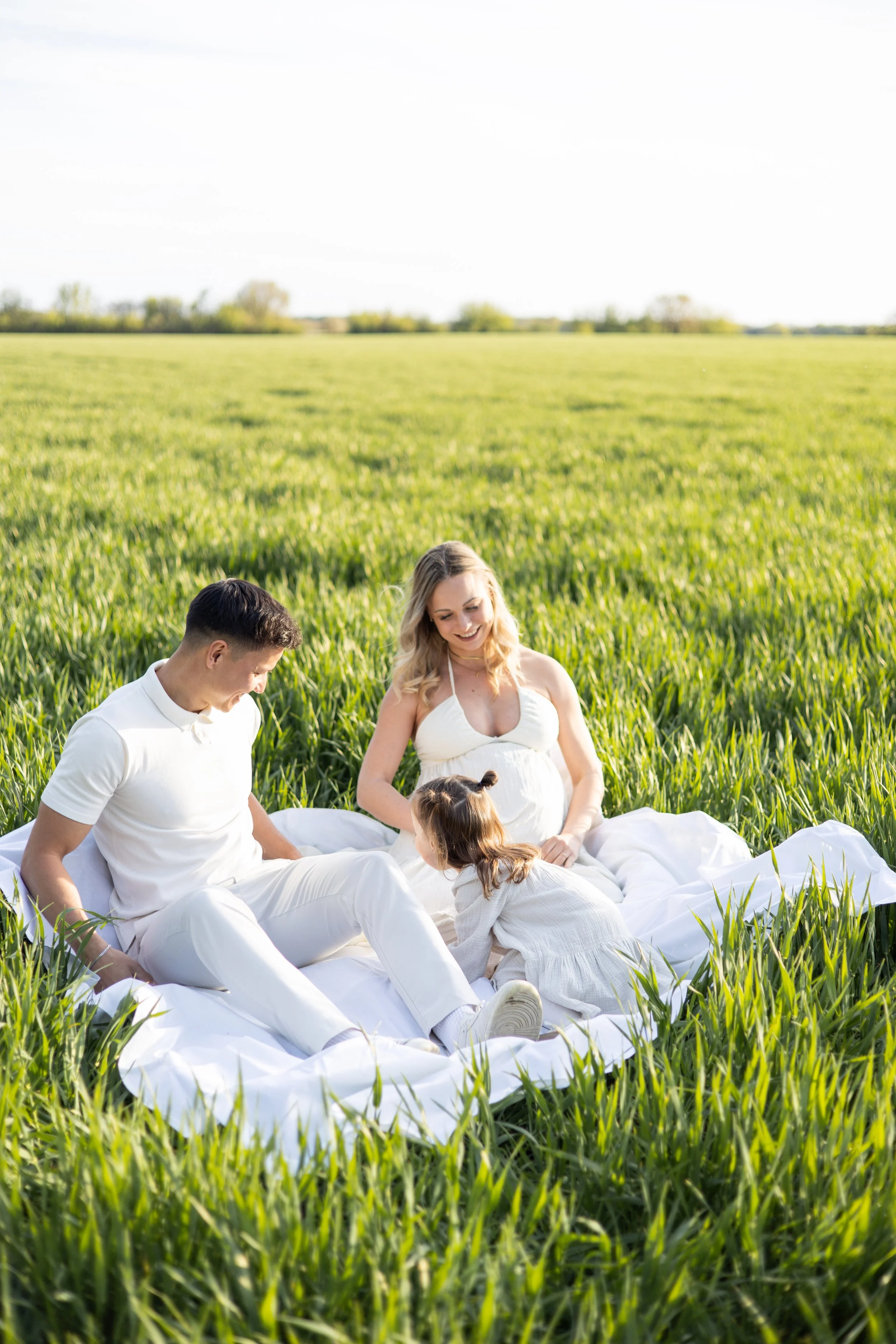 Eine Gruppe von vier Menschen, ein Mann, eine Frau, ein Mädchen und ein Baby, sitzen auf einer weißen Decke in einem grünen Feld bei Sonnenuntergang oder -aufgang und genießen die Natur.