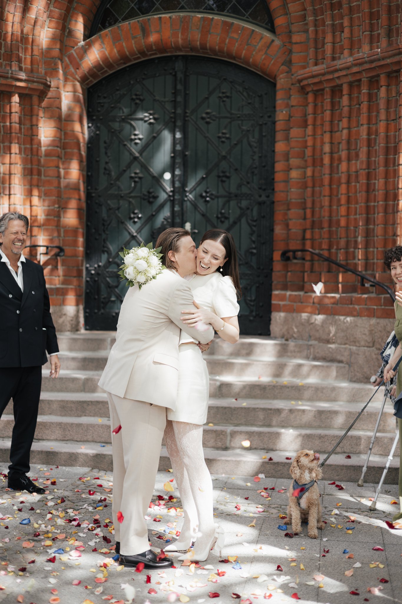 Ein Paar, das bei ihrer Hochzeit herzlich umarmt und küsst, steht vor einer roten Backstein- Kirche. Es ist sonnig, das Paar trägt elegante weiße Kleidung, und es sind Blumen und Konfetti auf den Stufen und auf dem Boden verteilt. Weitere Personen un