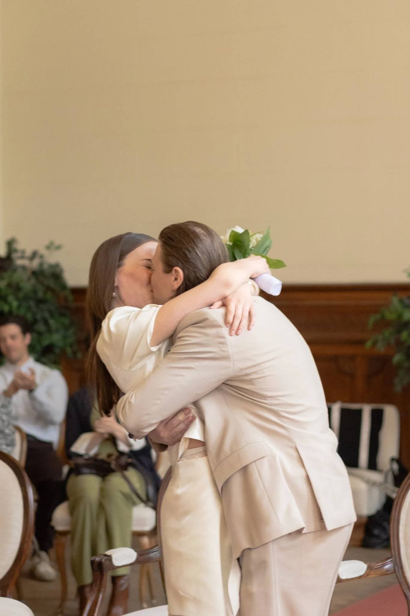 Ein Paar umarmt sich und küsst sich bei einer Hochzeit in einem Saal, im Hintergrund sitzen Gäste, die das Ereignis beobachten.