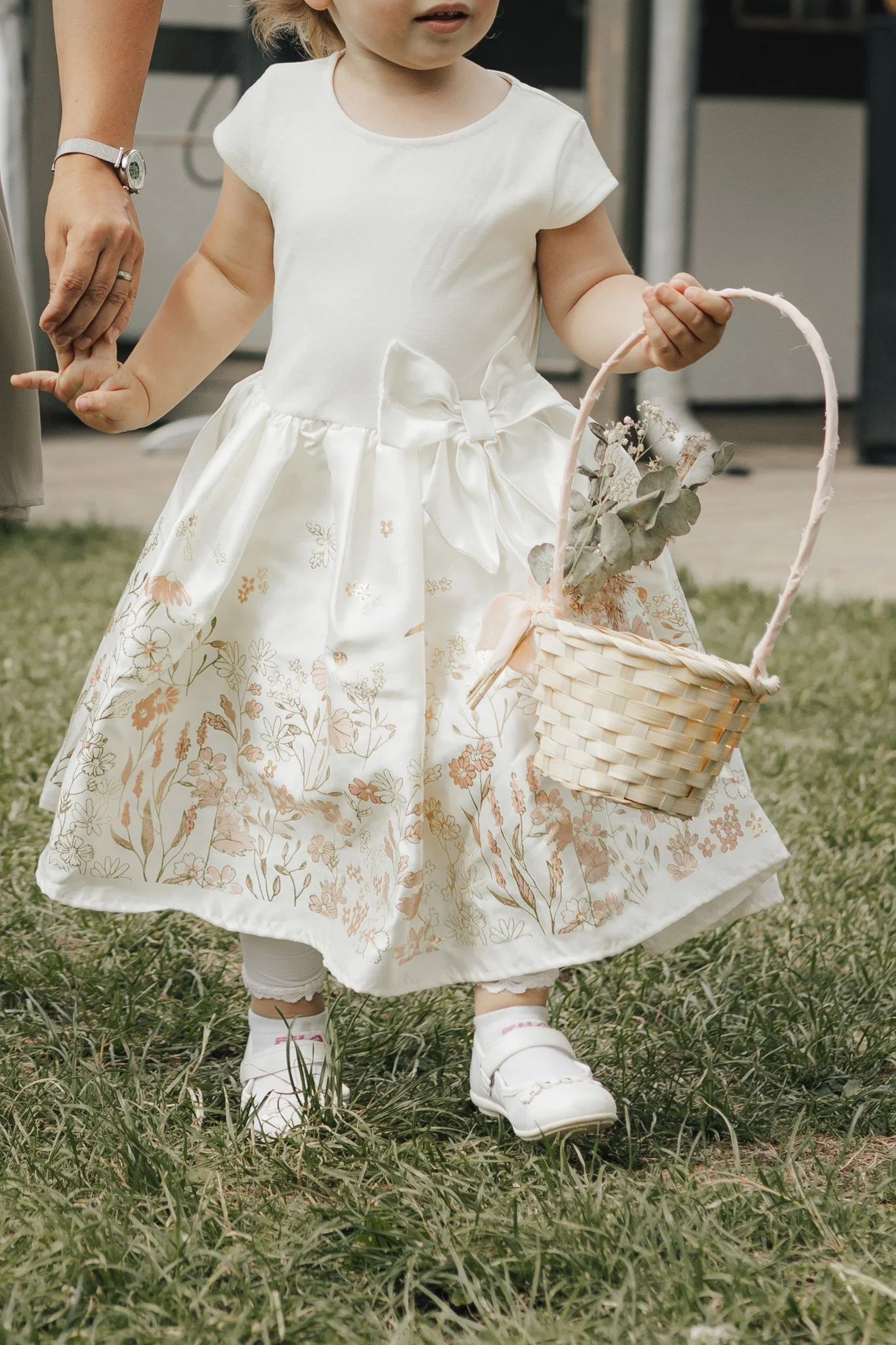 Ein kleines Mädchen trägt ein weißes Kleid mit Blumenmuster, weiße Schuhe und hält einen Korb mit Blättern. Es steht auf einer Wiese.