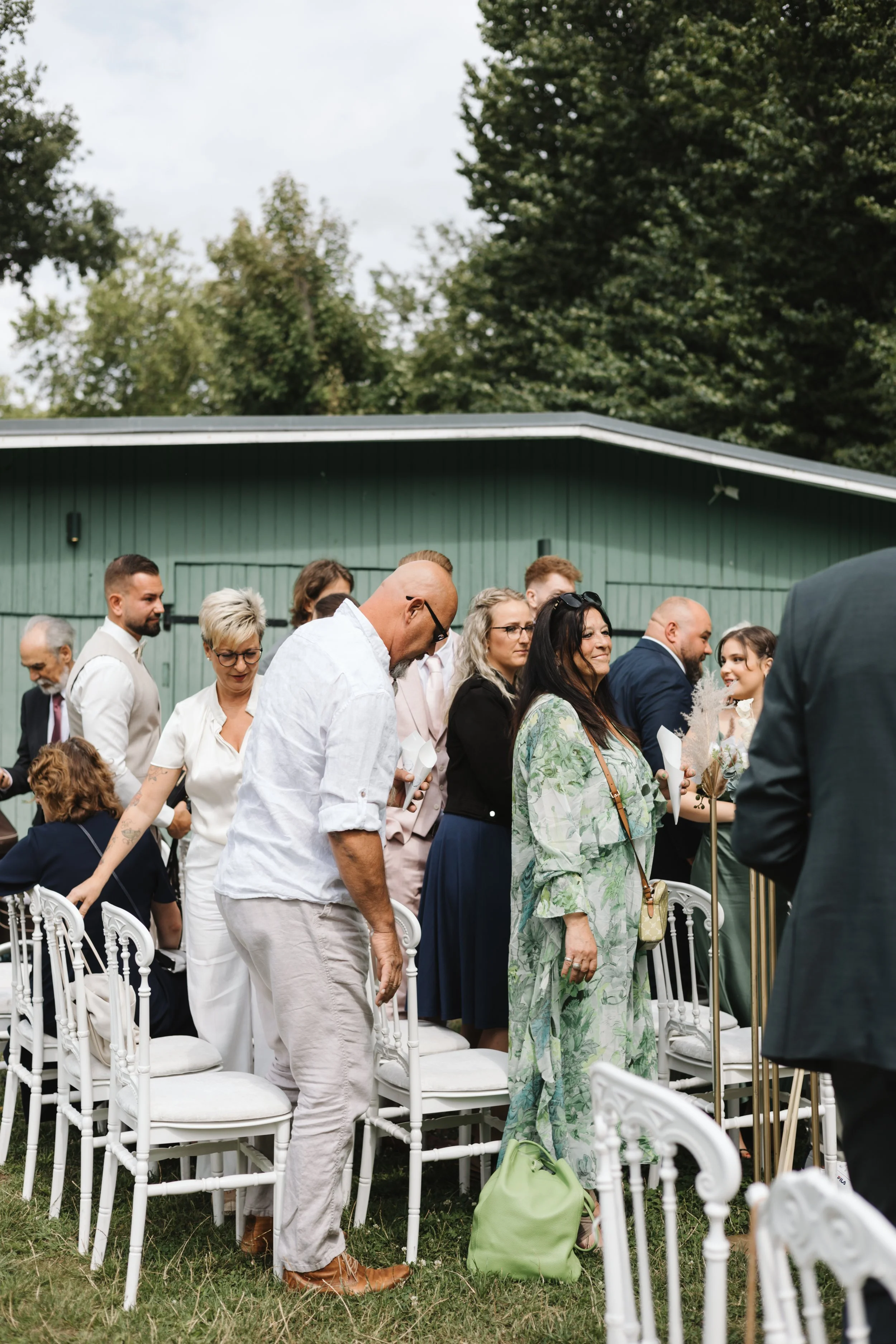 Eine Gruppe von Menschen steht bei einer Outdoor-Hochzeit, einige mit Blick nach vorne, andere mit Blick nach unten oder zur Seite, auf weißen Stühlen auf grünem Rasen, im Hintergrund grüne Bäume und ein graues Gebäude.