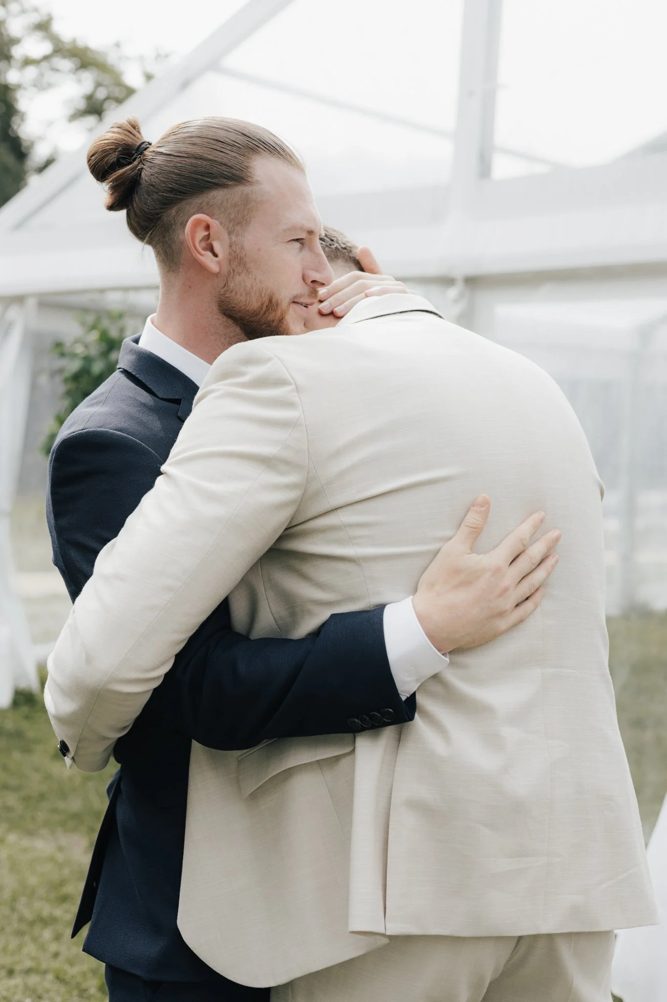 Zwei Männer umarmen sich herzlich, einer trägt einen dunklen Anzug, der andere einen hellen Anzug, im Außenbereich, wahrscheinlich bei einer Hochzeit oder Feier.