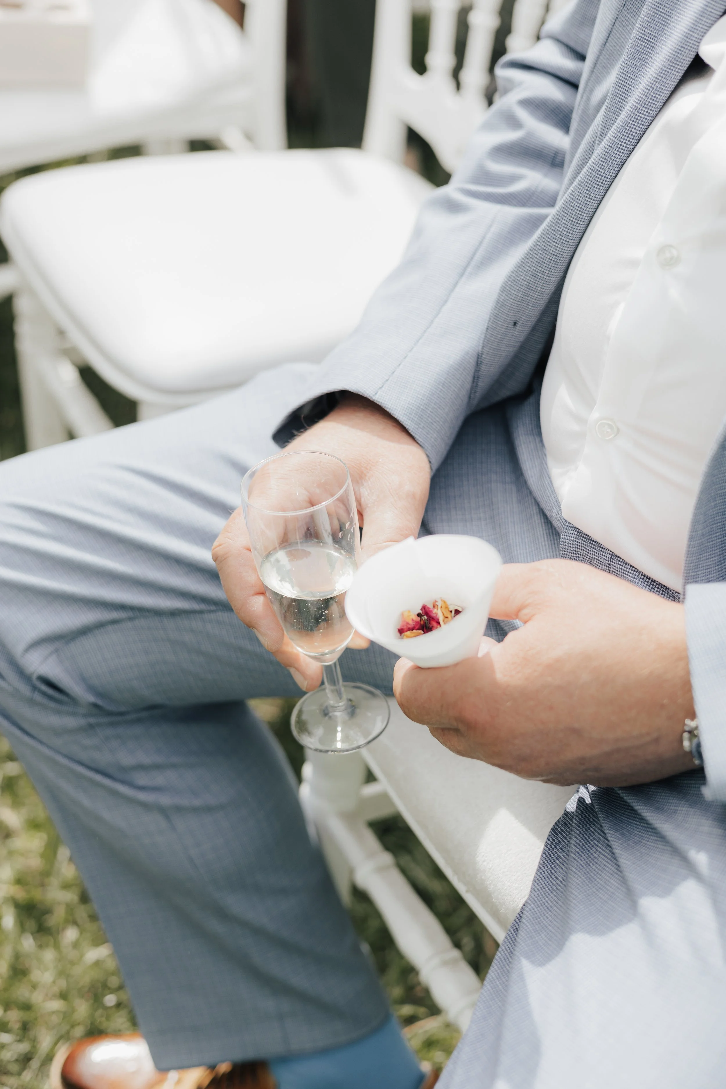 Nahaufnahme von einem Mann in einem grauen Anzug, der ein Gläschen Sekt hält, während eine andere Person ihm aus einer weißen Schale mit Blumenblüten etwas serviert. Es scheint sich um eine festliche Gelegenheit im Freien zu handeln.