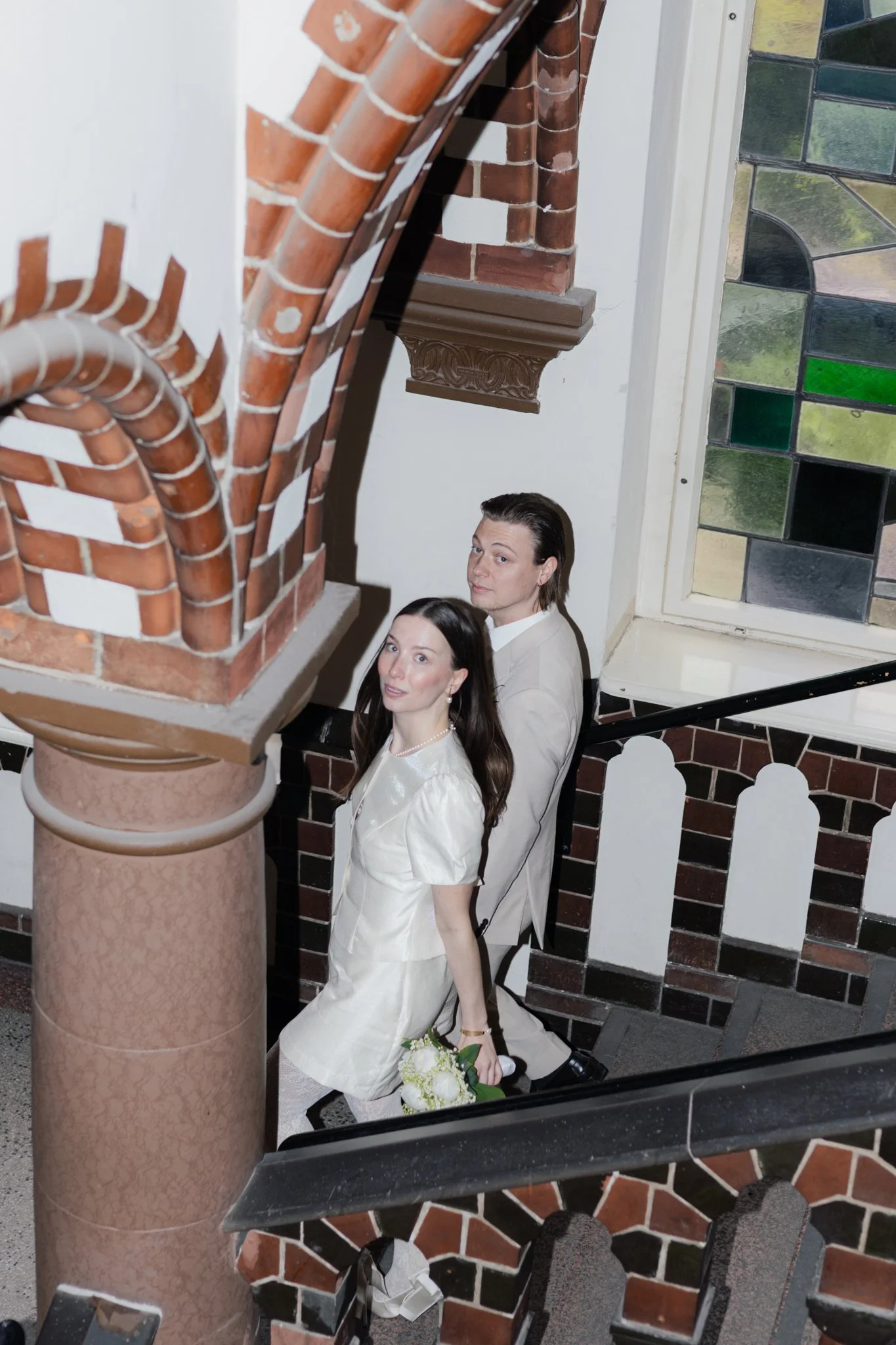 Zwei Hochzeitsgäste, ein Mann und eine Frau, gehen die Treppe im öffentlichen Gebäude hinauf. Die Frau trägt ein weißes Hochzeitskleid und hält einen Blumenstrauß. Der Mann trägt einen hellen Anzug. Das Gebäude hat bunte Glasscheiben im Fenster und e