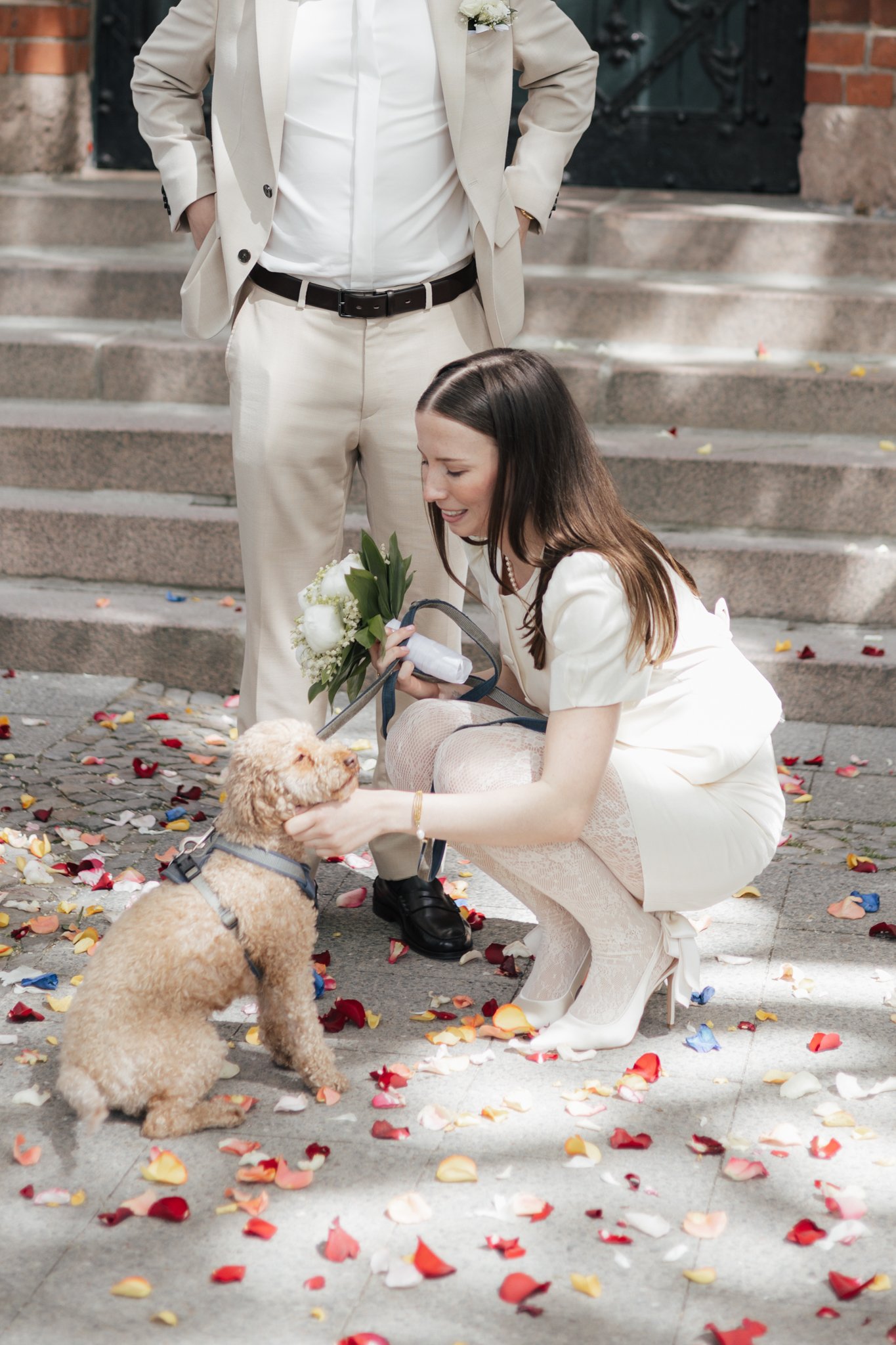 Ein Brautpaar bei einer Hochzeit mit einem Hund auf den Treppen vor einem schwarzen Tür im Freien, mit Blumen und Blütenblättern auf dem Boden.