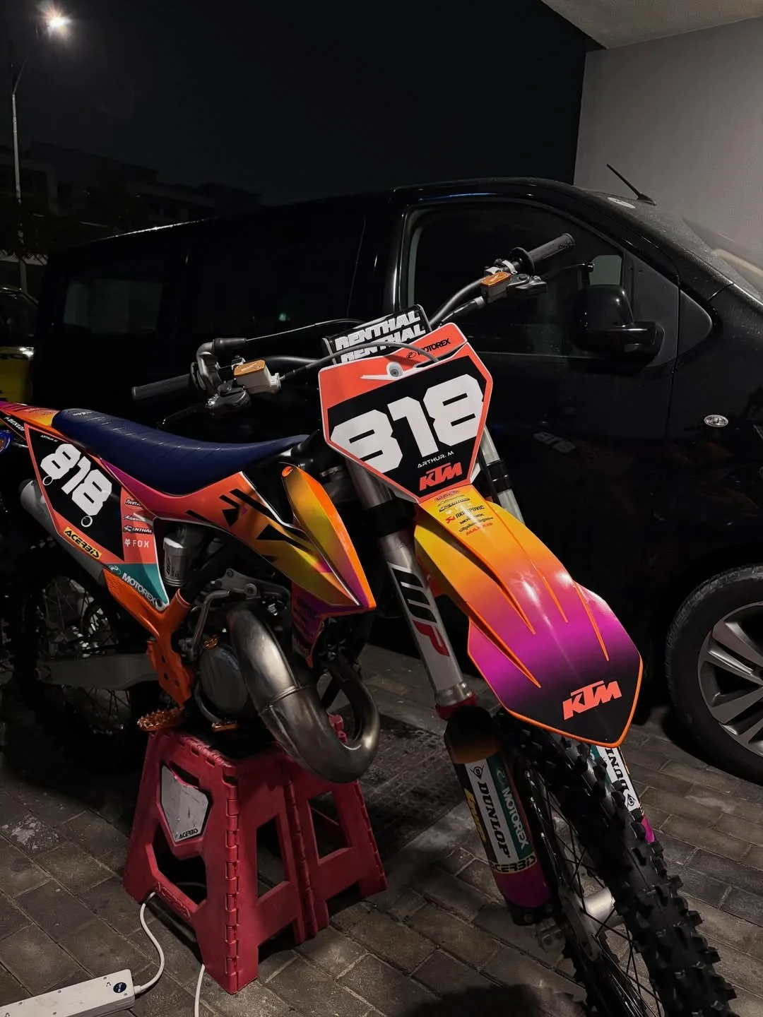 A KTM dirt bike with a colorful orange, pink, and purple design, parked on a small red stool in front of a dark-colored car at night.