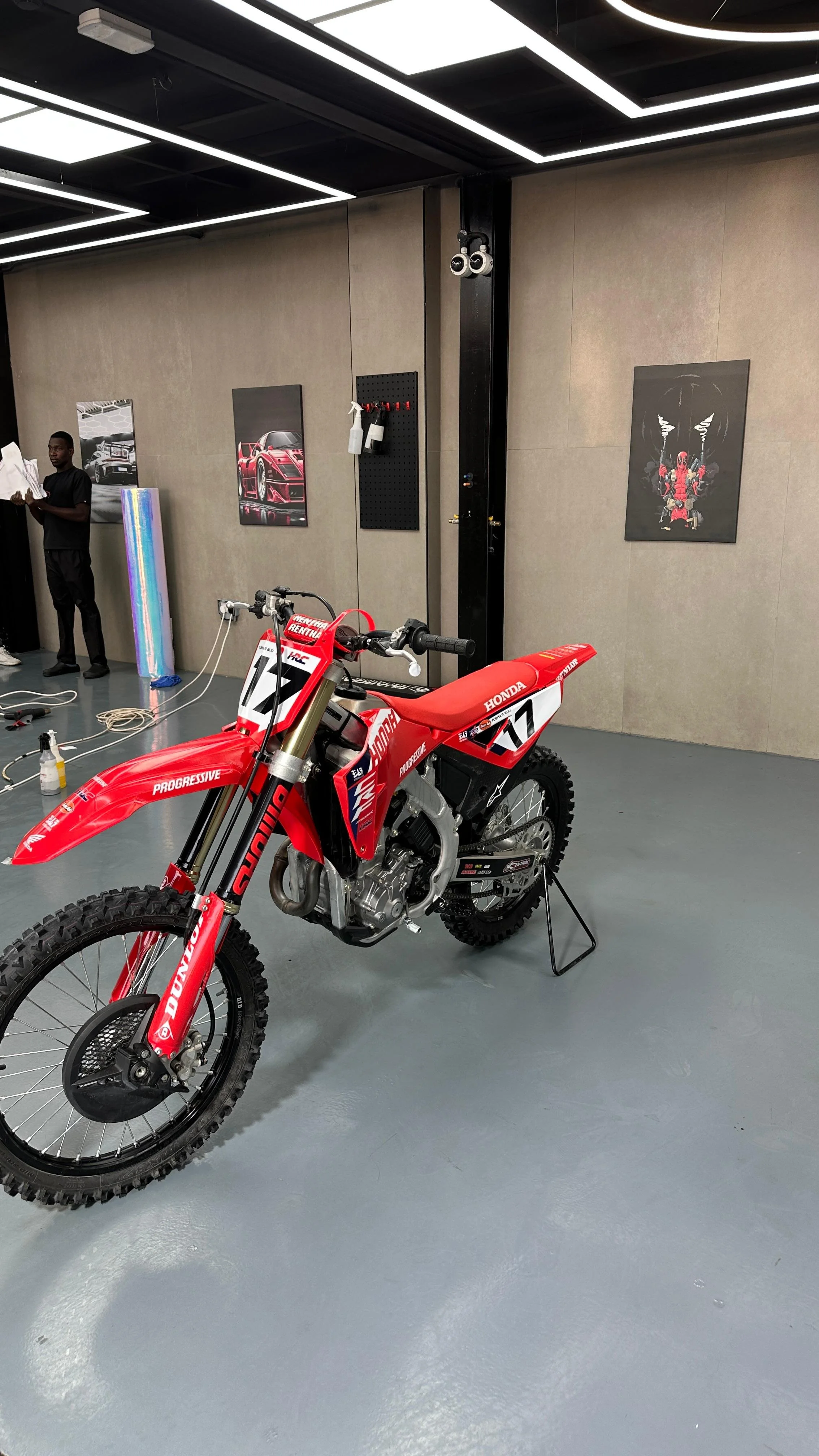 Red Honda dirt bike with racing number 77 in a showroom or exhibition space, with framed automotive and art posters on the walls.