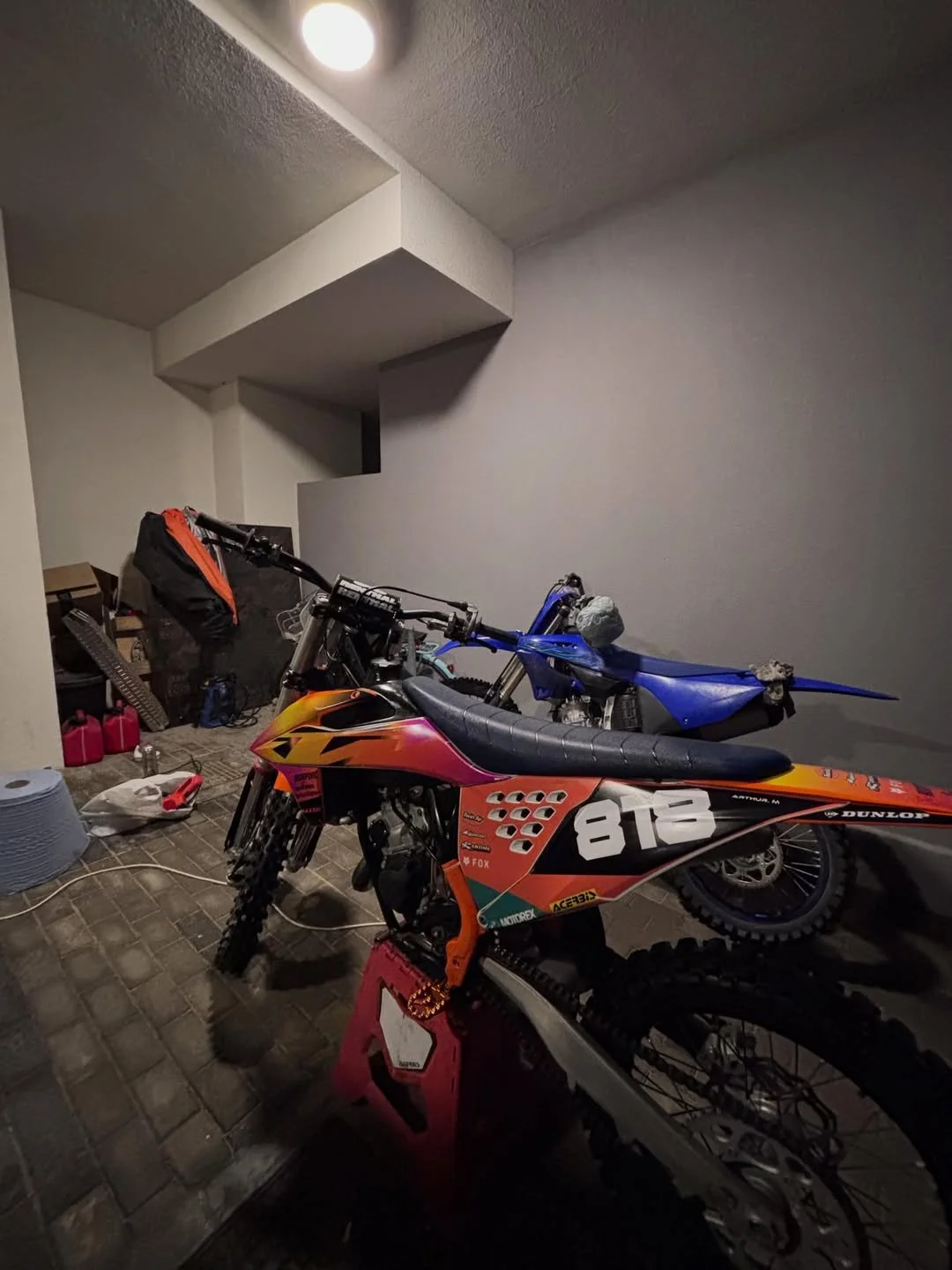 Indoor parking area with a dirt bike, motorcycle, and a blue scooter parked. There are various items on the floor and a large gray container.
