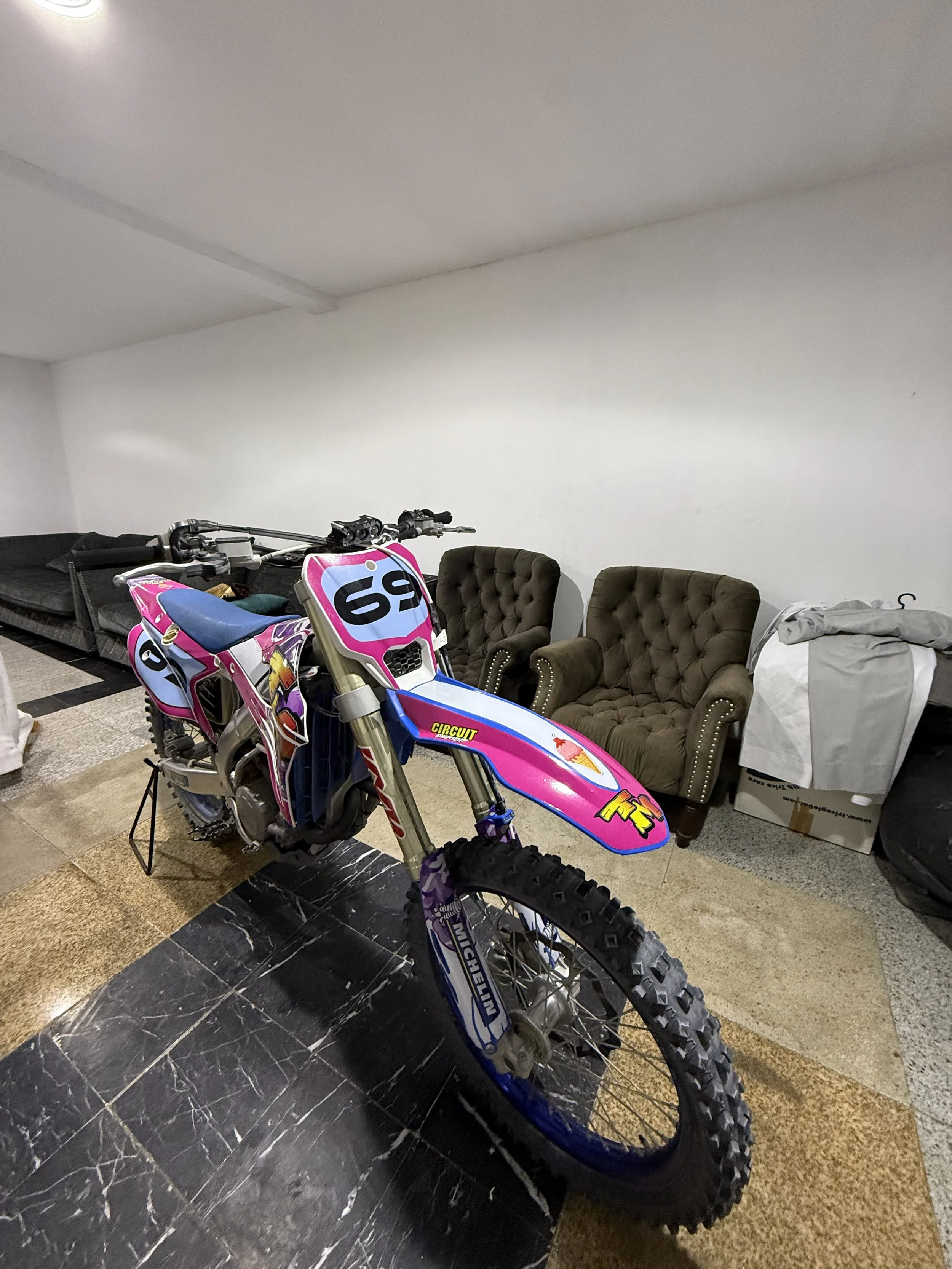 Indoor scene with a pink, purple, and blue motocross bike in the foreground, chairs and boxes in the background, and a white wall.