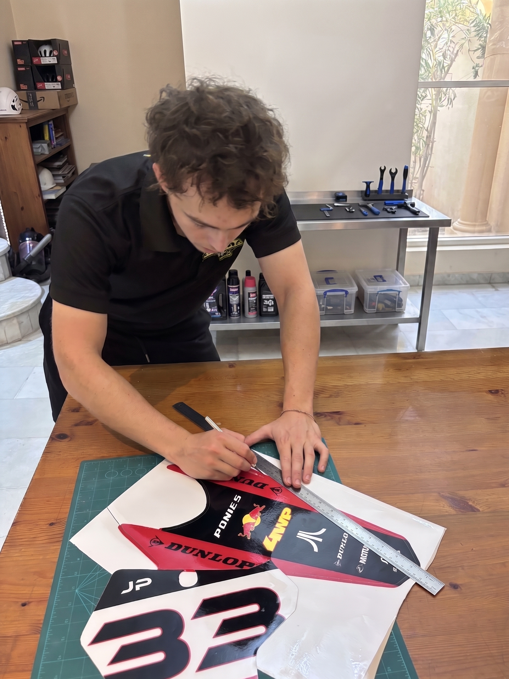A person is measuring a large racing decal on a wooden table using a metal ruler, with tools and decals visible in the background.