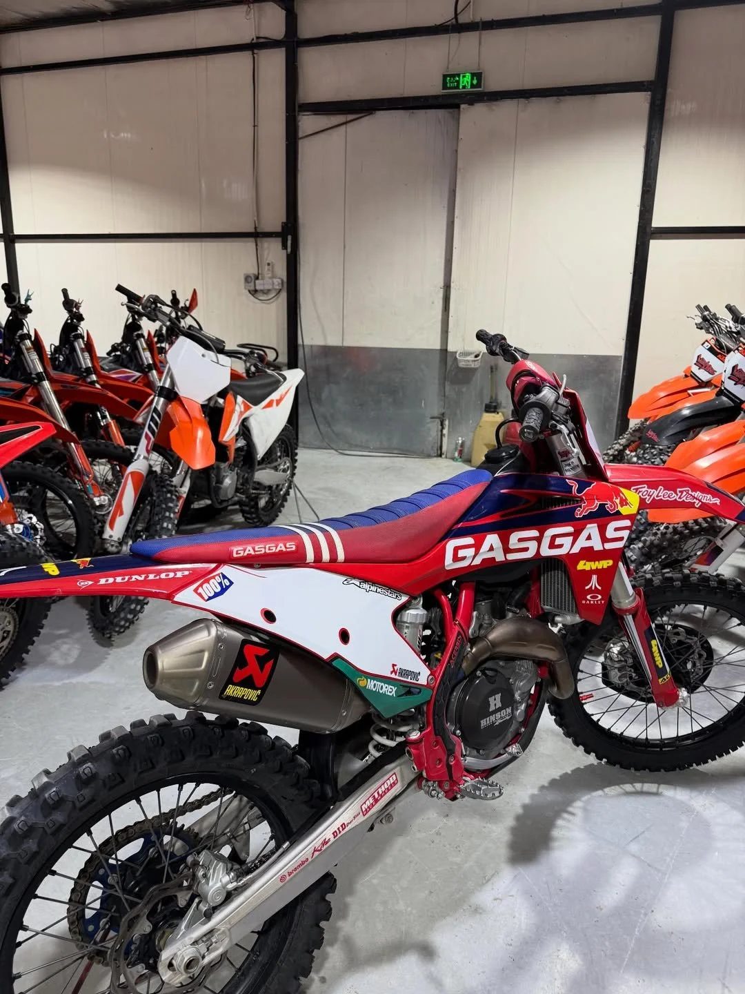 A collection of motocross bikes in a showroom or storage area, with the prominent bike in the foreground featuring red, blue, and white racing decals and branding, along with a metallic exhaust pipe and knobby tires.