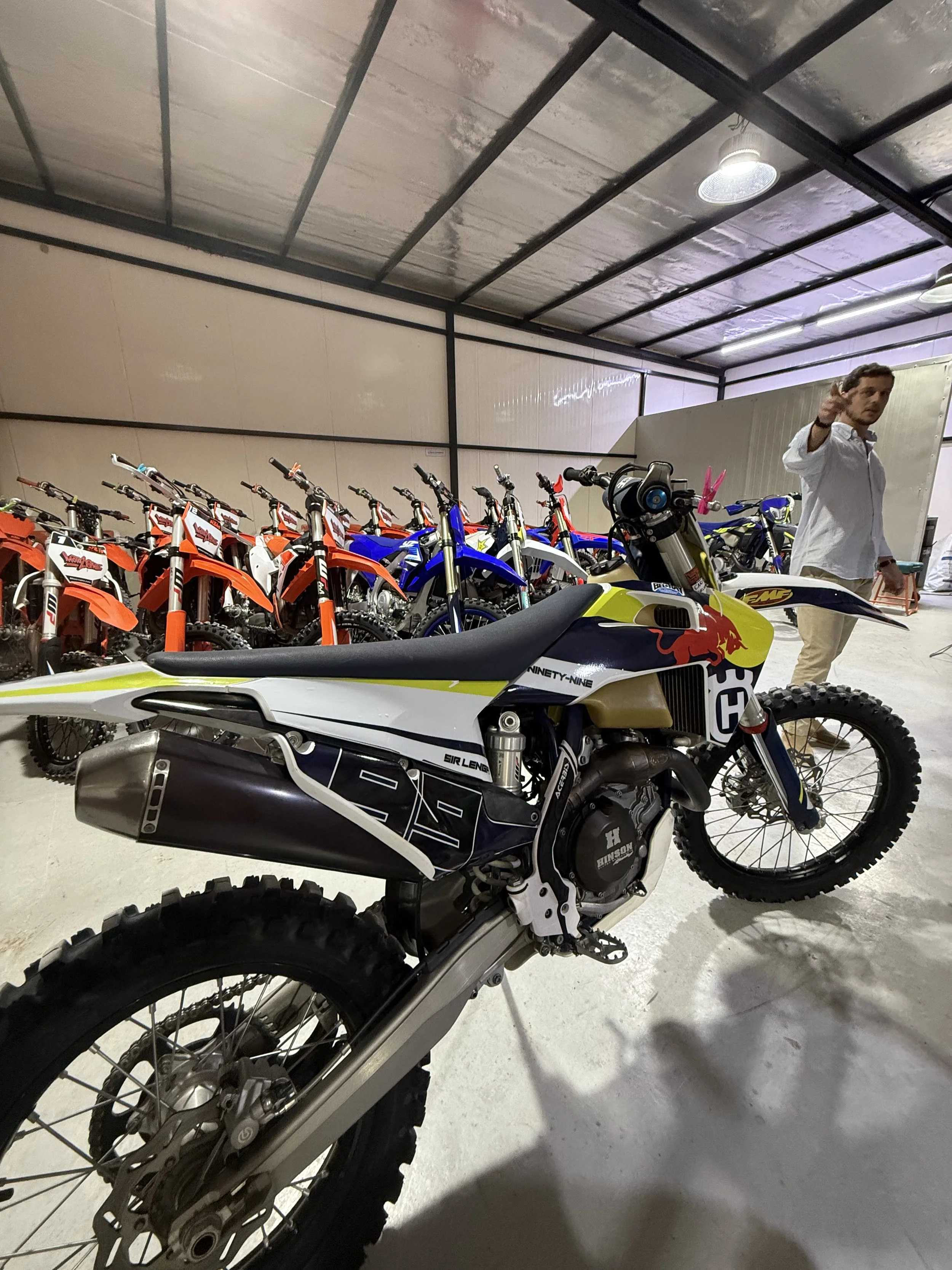 An indoor area with multiple dirt bikes parked in a row, including models with vibrant colors such as orange, blue, and white. A man in a white shirt and beige pants is standing nearby, pointing or gesturing.