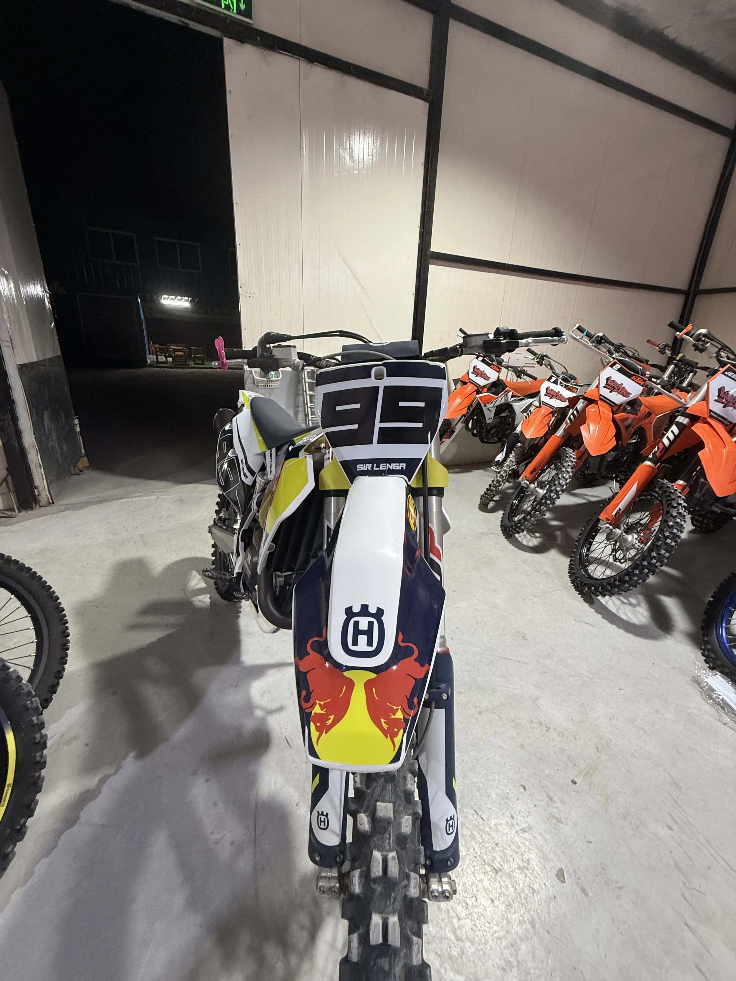 From the front, a motocross bike with a Red Bull logo on the front fender, surrounded by several orange dirt bikes in a garage or storage area with a concrete floor and white and black walls.