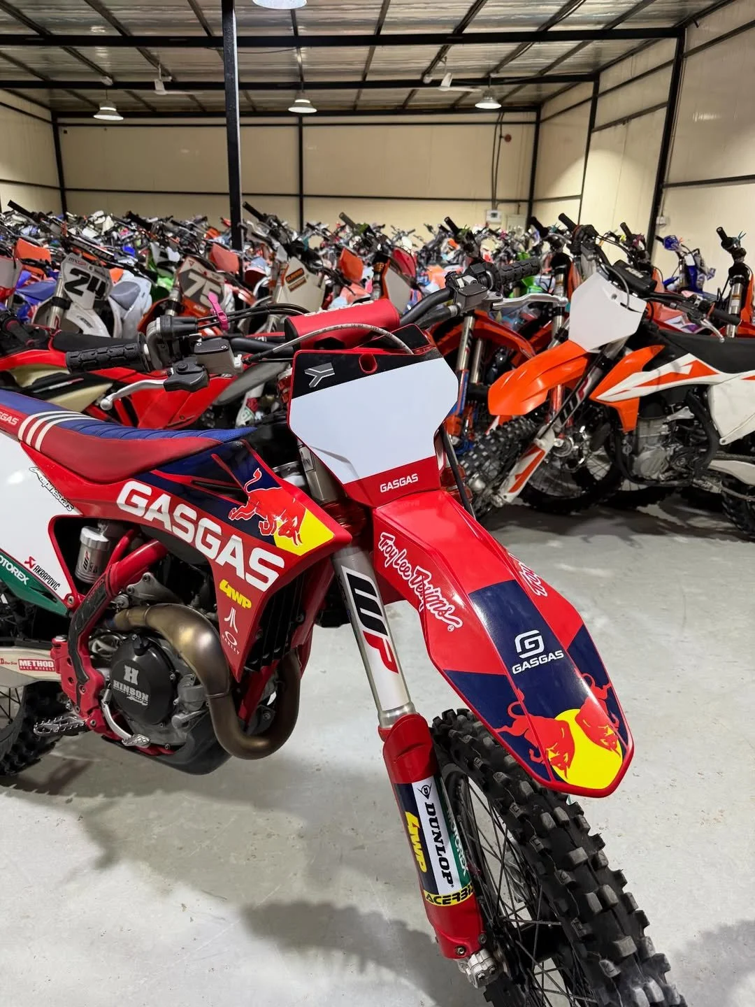 Red and blue GasGas motocross dirt bike in front of a large collection of bikes inside a warehouse or storage building.