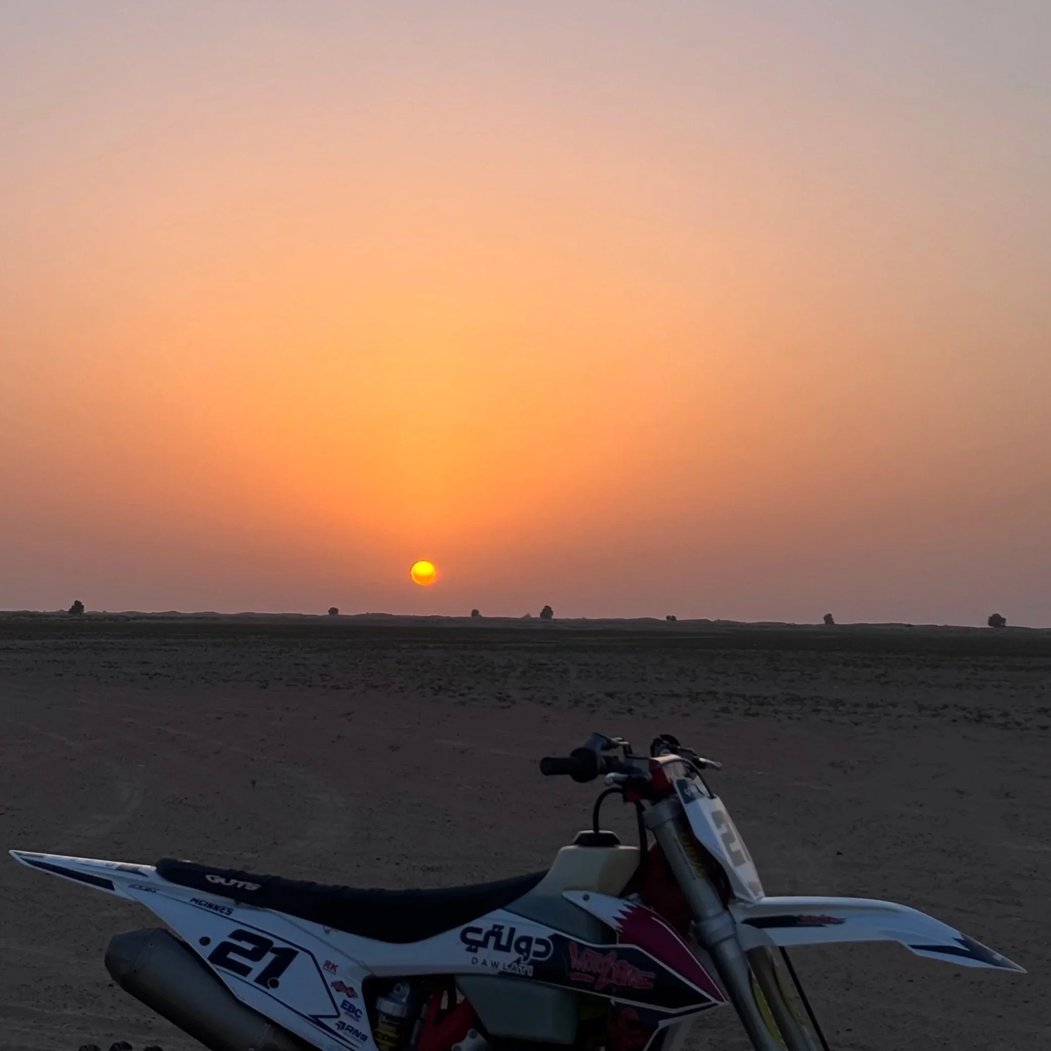A dirt bike parked on a sandy terrain with a sunset or sunrise in the background. The sky is a gradient of soft pink and peach colors, and the sun is near the horizon with a small group of trees in the distance. The bike appears to have racing decals