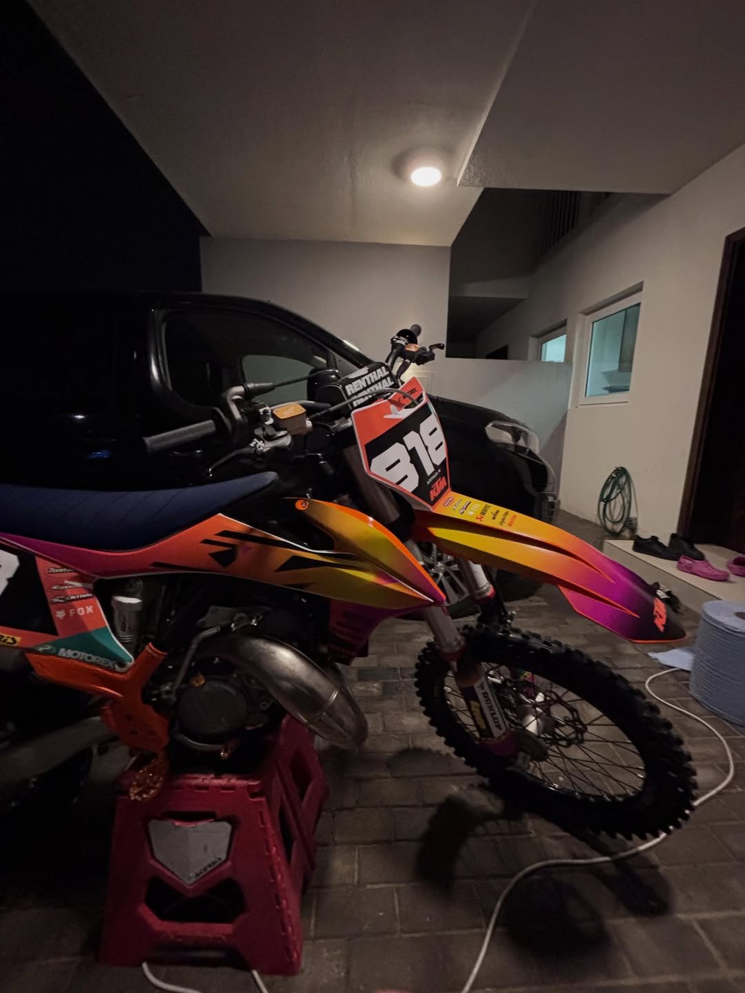 A dirt bike with orange, pink, and yellow colors set inside a garage next to a black car, with a pink stand under the bike's engine and various household items visible in the background.