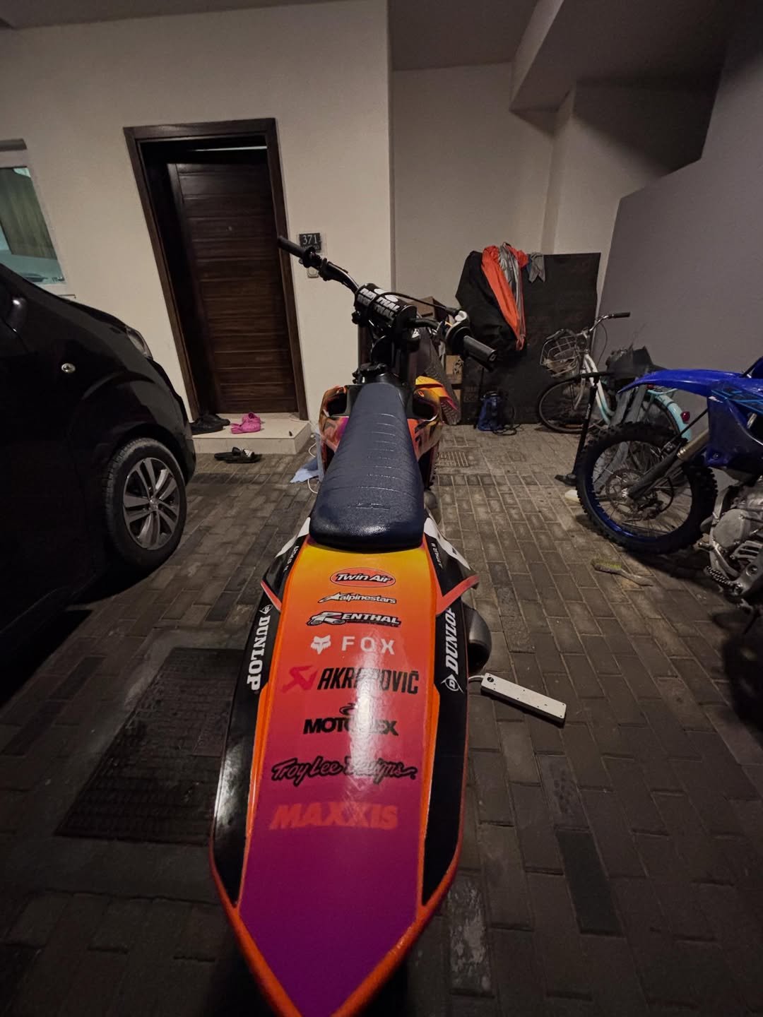 A racing motorcycle parked in a driveway, viewed from the front, with a car on the left side and a bicycle and motorcycle on the right side, next to a house entrance with the number 371.