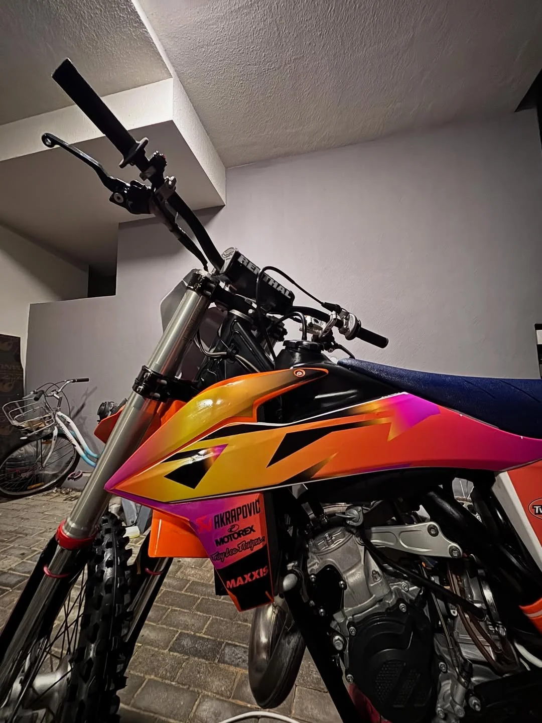 Close-up of a brightly colored motocross motorcycle with orange, pink, and yellow graphics, parked indoors on a brick floor, with another bicycle visible in the background.
