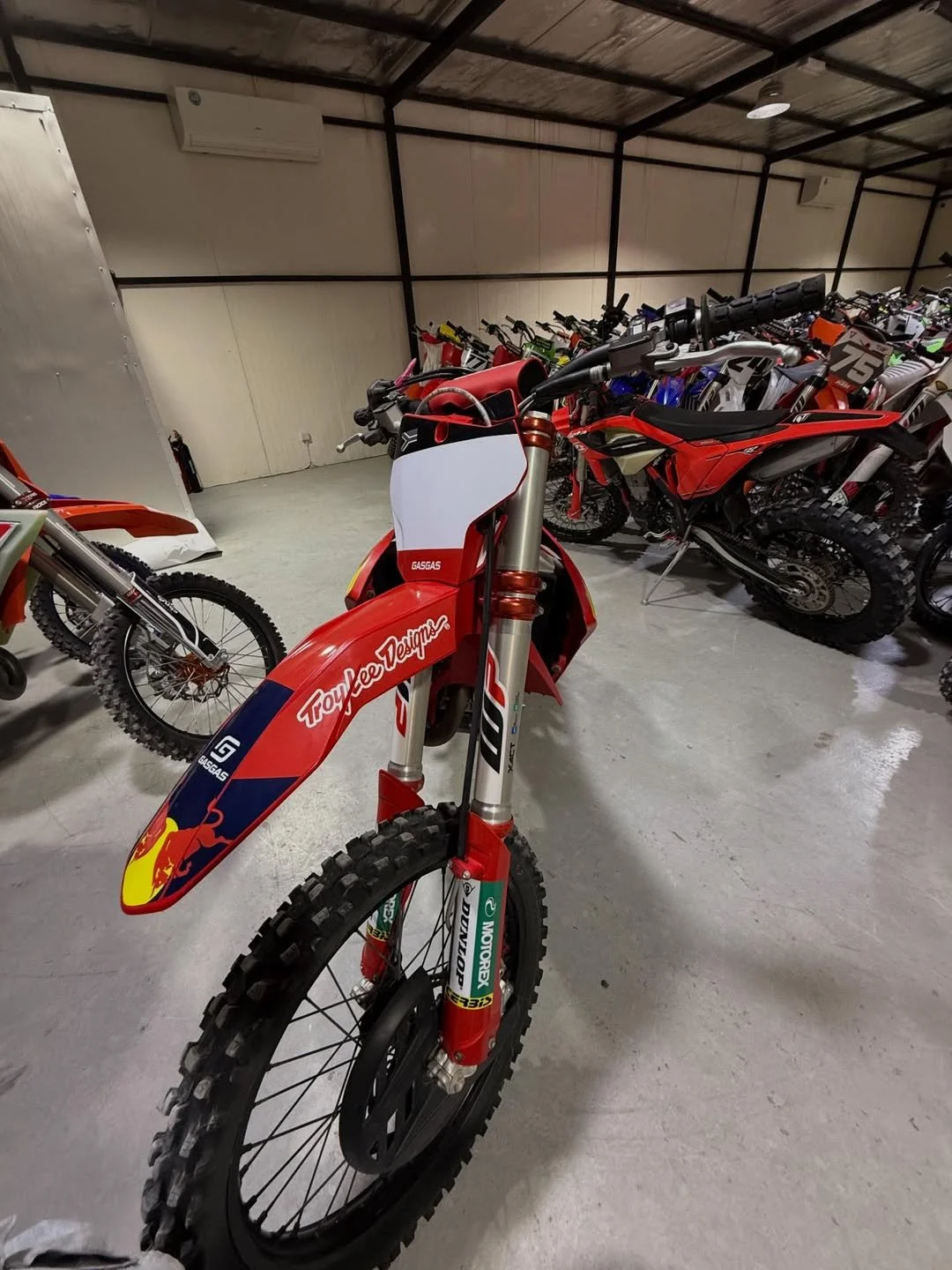 A red and white motocross dirt bike with 'Troy Lee Designs' on the fender, surrounded by other motorcycles in a warehouse or showroom.