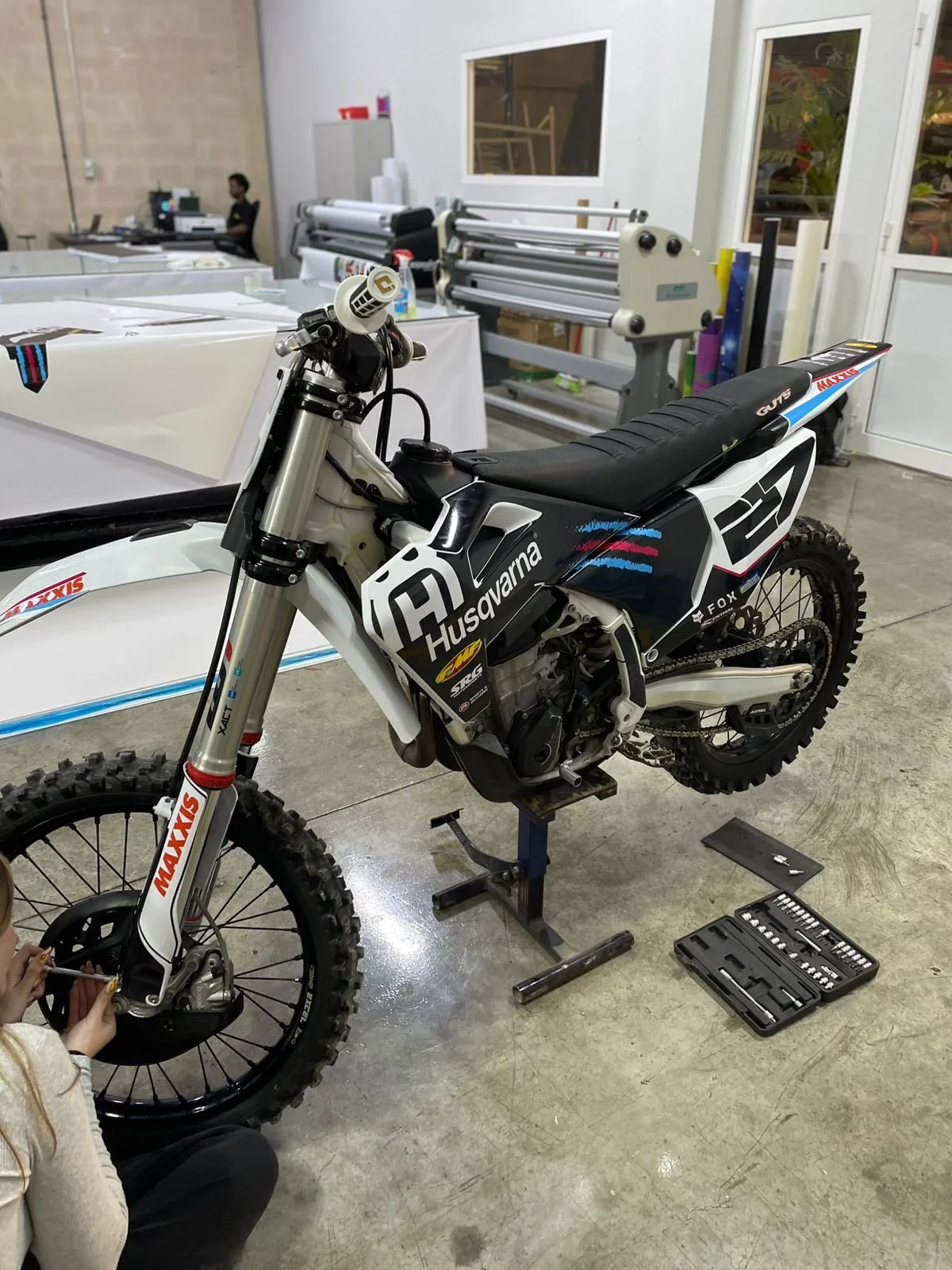 A Husqvarna dirt bike is being repaired or assembled in a workshop. A person is working on the front wheel with tools, and there is a toolkit on the floor. The bike has Maxxis tires and Husqvarna branding, with a black, white, and blue livery. The ba