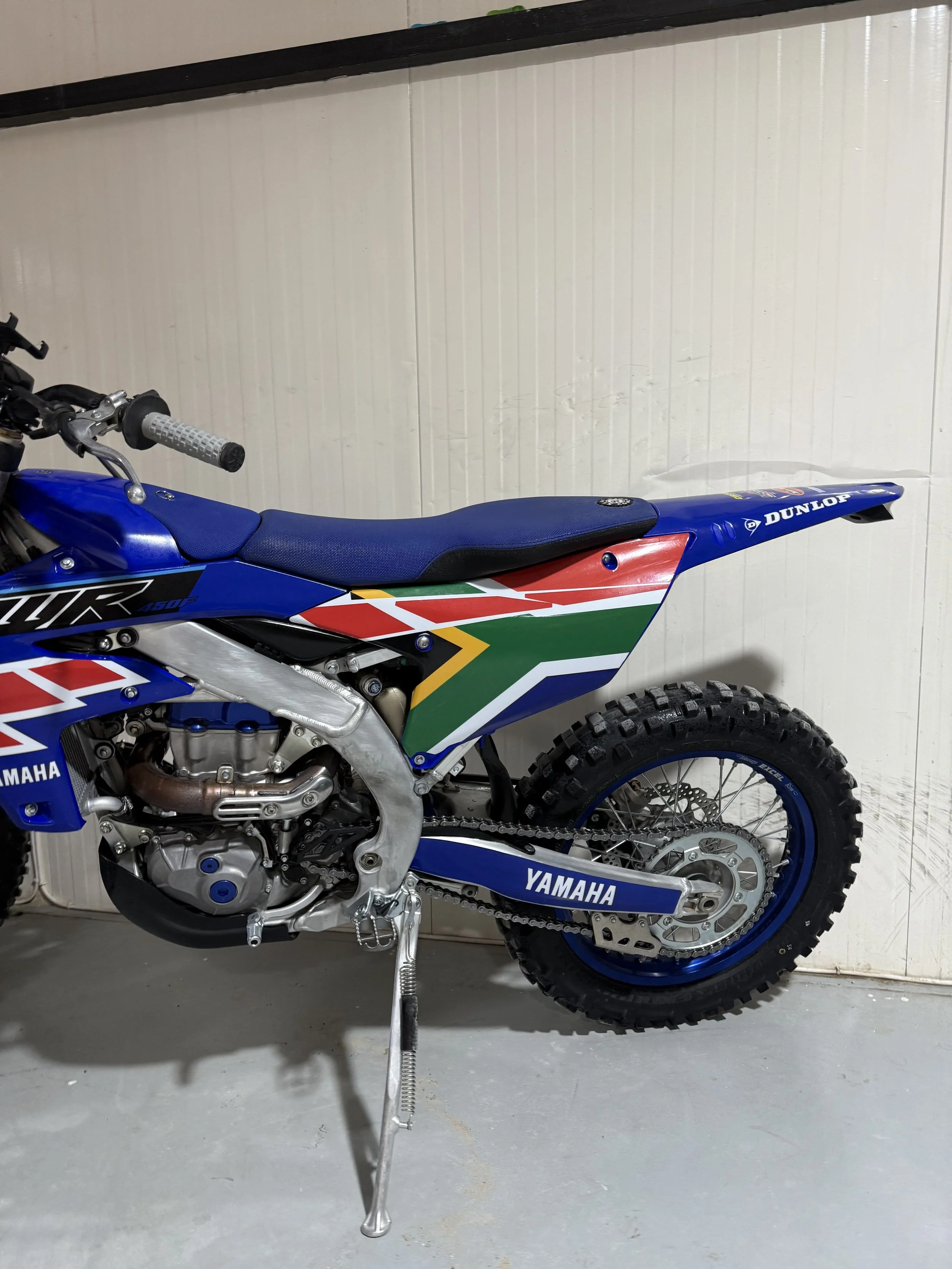 A blue Yamaha dirt bike with racing graphics is parked in a garage, supported by a kickstand. The bike features knobby tires, a racing seat, and branding decals including Dunlop and Yamaha.