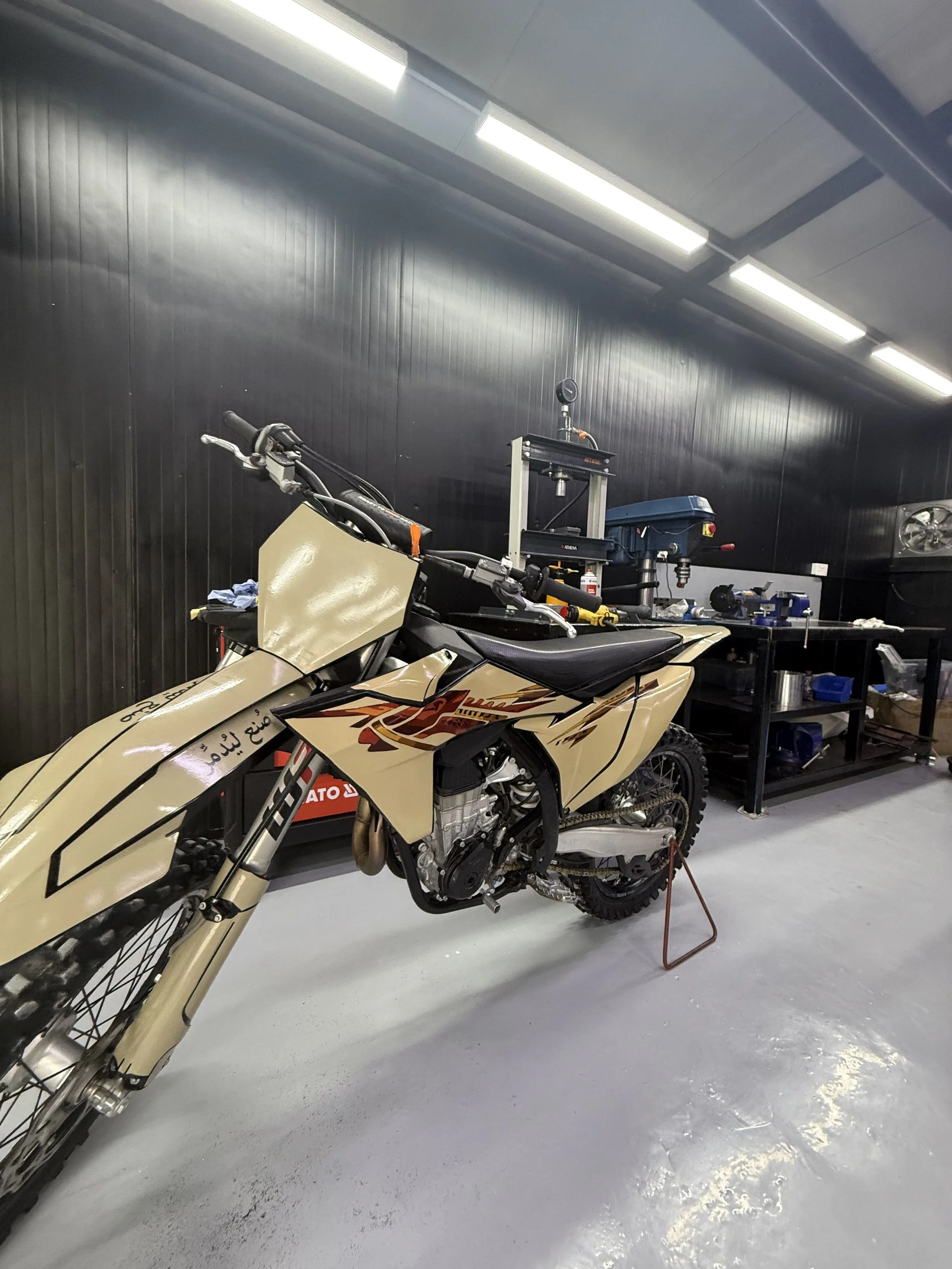 A beige off-road motorcycle is in a garage or workshop. The motorcycle is partially disassembled, with some parts removed. The background includes a black wall, fluorescent lights on the ceiling, and a workbench with tools and equipment.