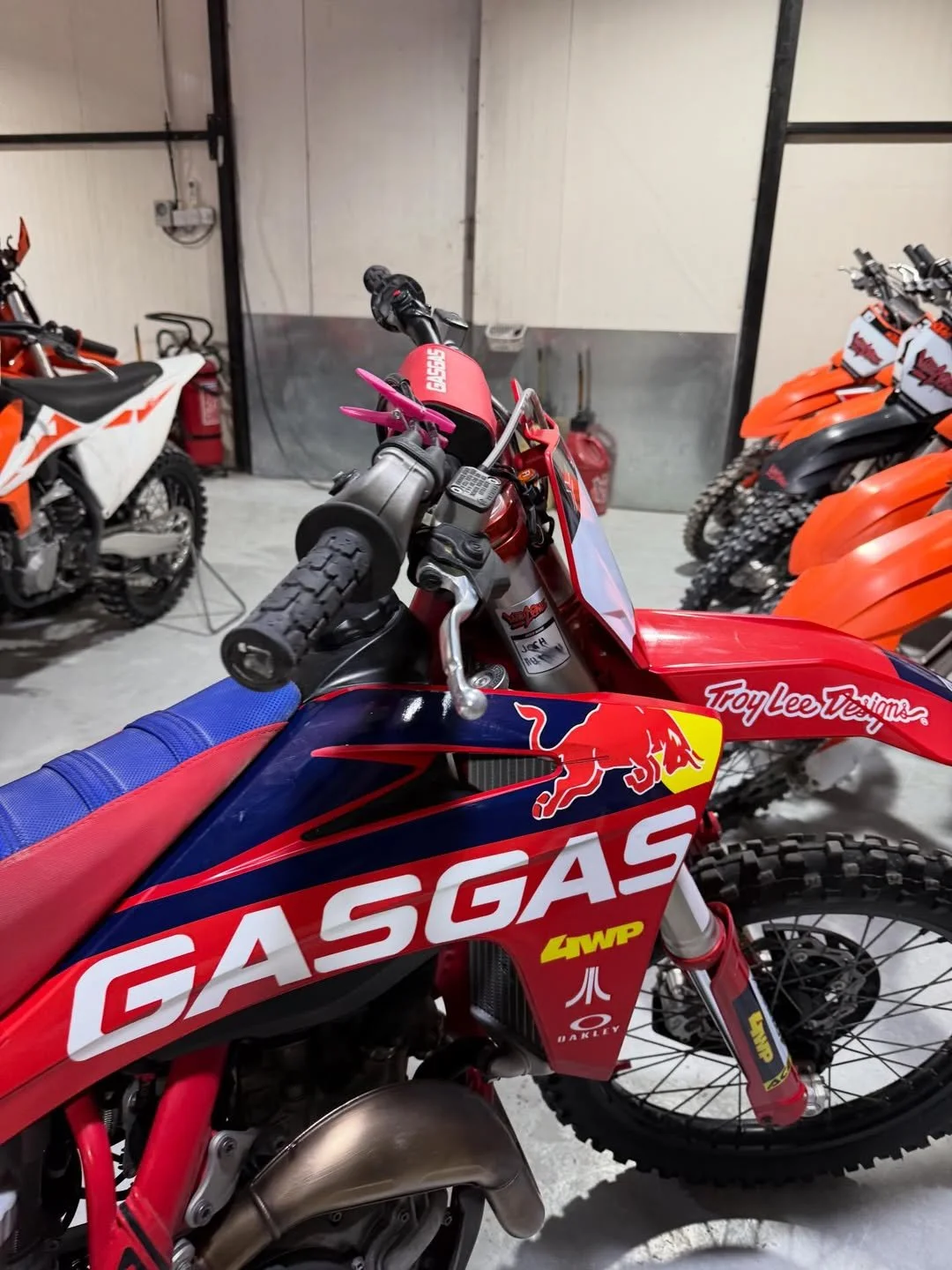 Close-up of a red and blue GasGas dirt bike with Troy Lee Designs branding, surrounded by other dirt bikes in a garage or workshop setting.