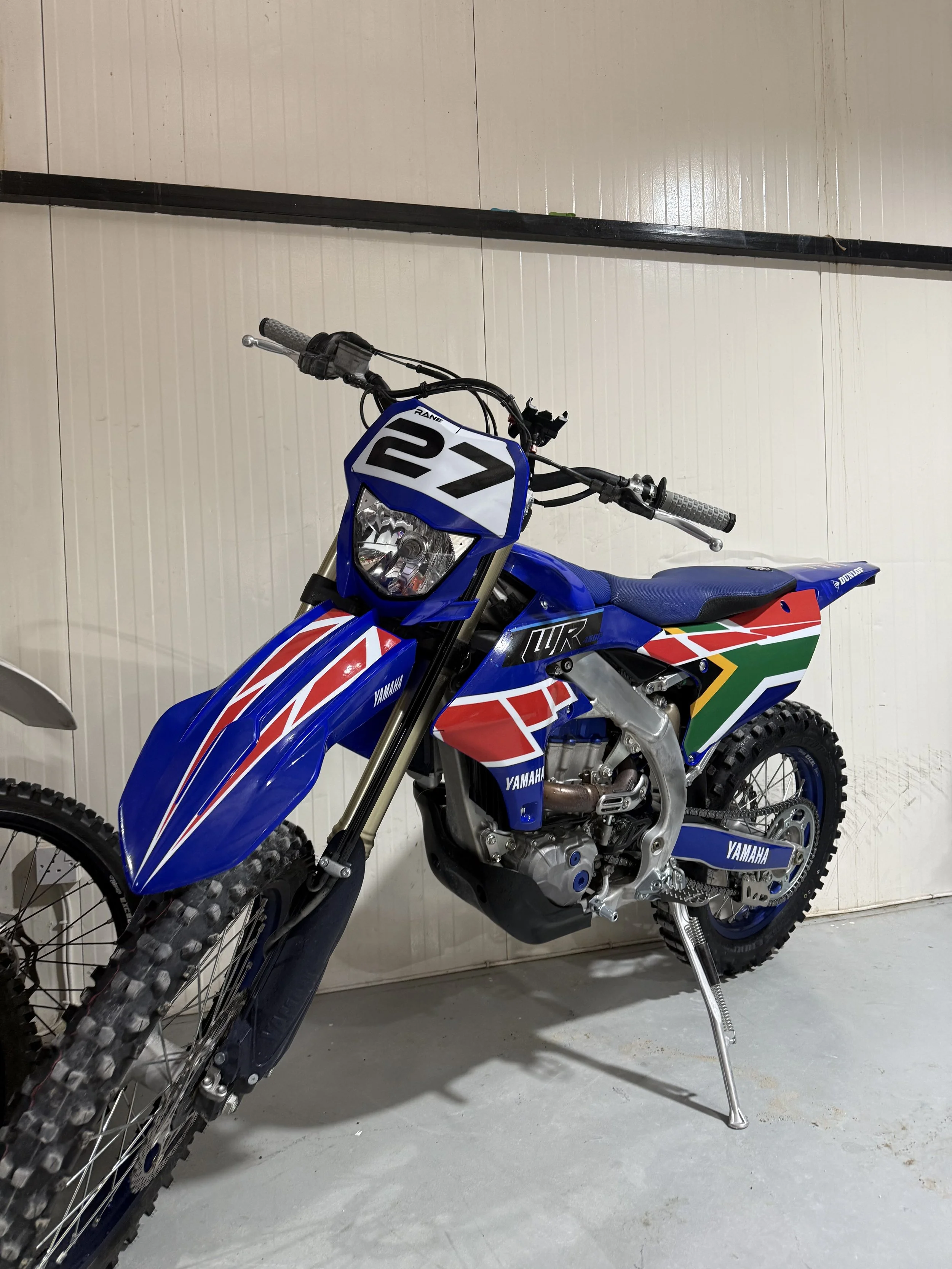 A blue Yamaha dirt bike with racing number 21 on the front, positioned indoors against a beige wall.