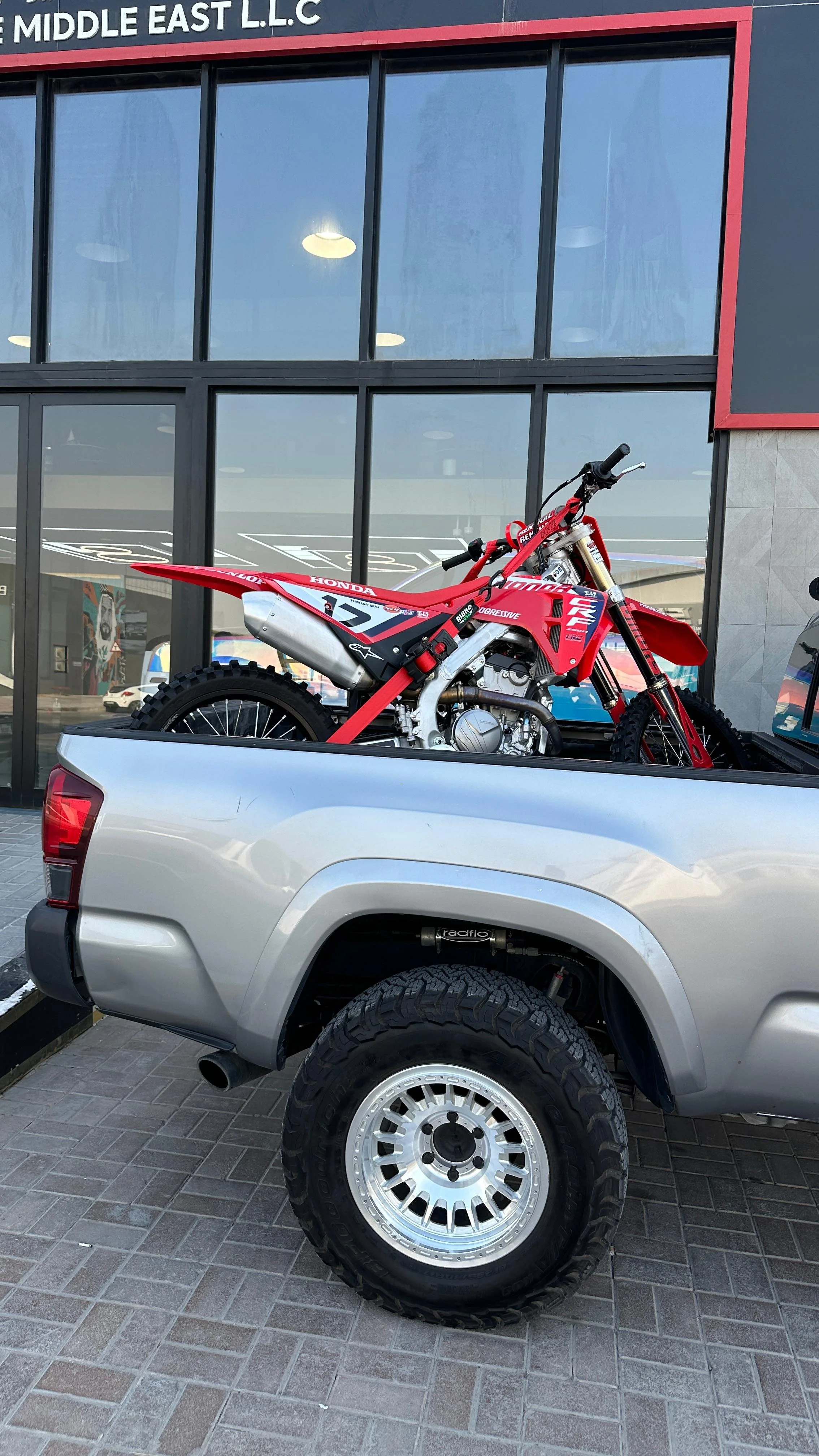 A red Honda dirt bike is loaded onto the back of a silver pickup truck parked outside a modern building with large glass windows.