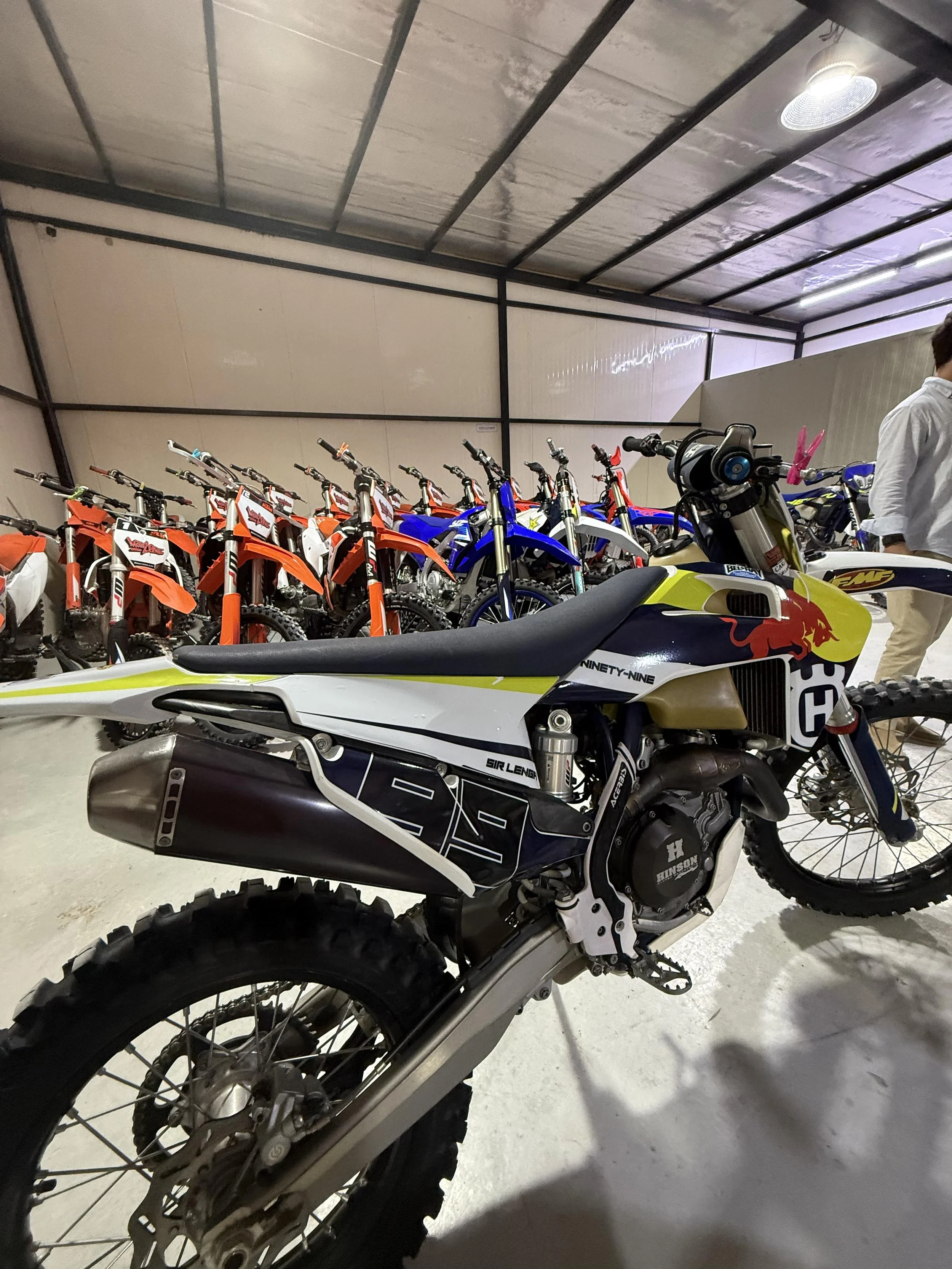 A garage with multiple dirt bikes lined up, including a yellow and black dirt bike in the foreground and several orange and blue bikes in the background.