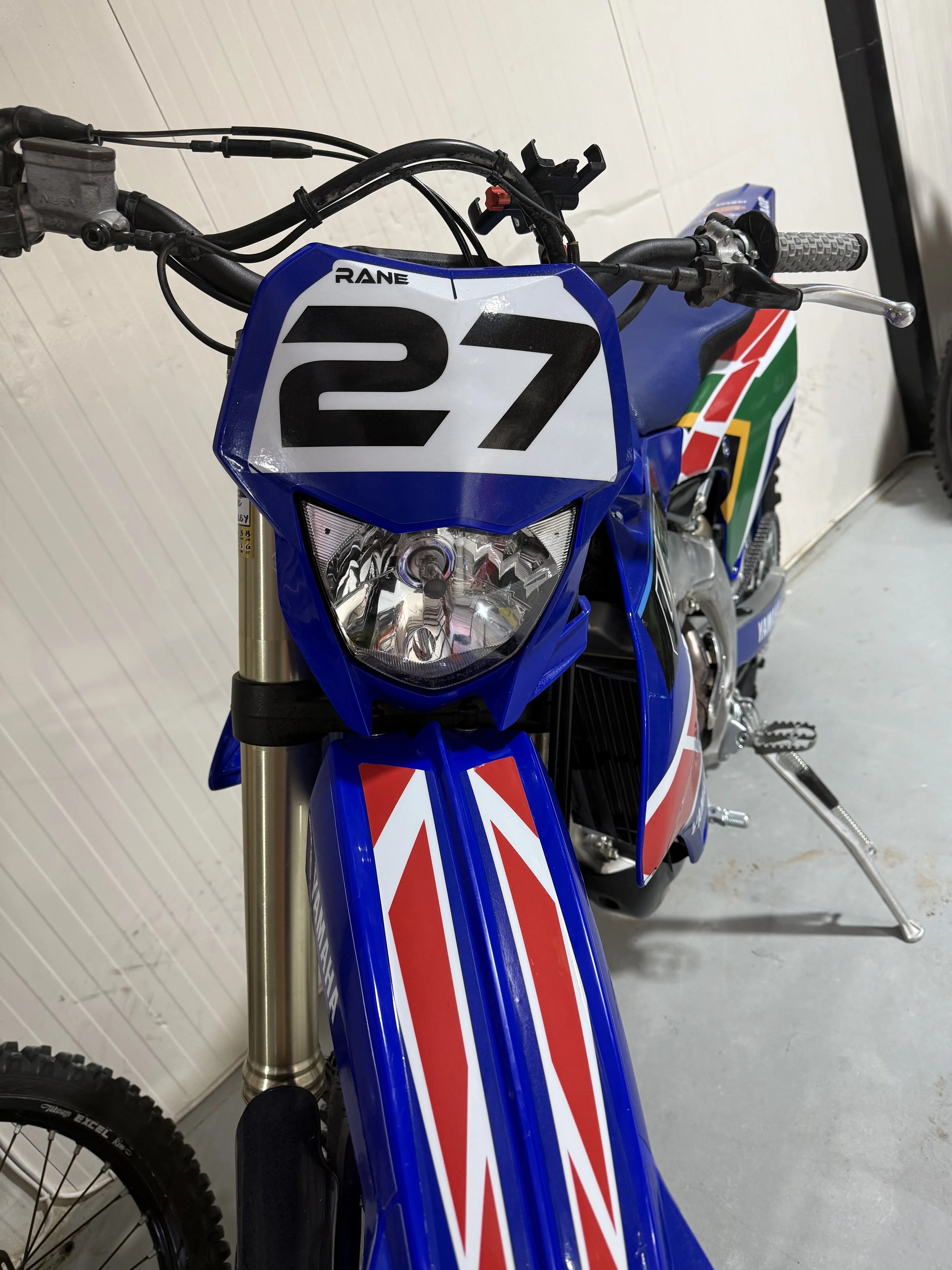 A blue and white dirt bike with a number 27 plate on the front, showing the handlebars, headlight, and part of the front wheel.