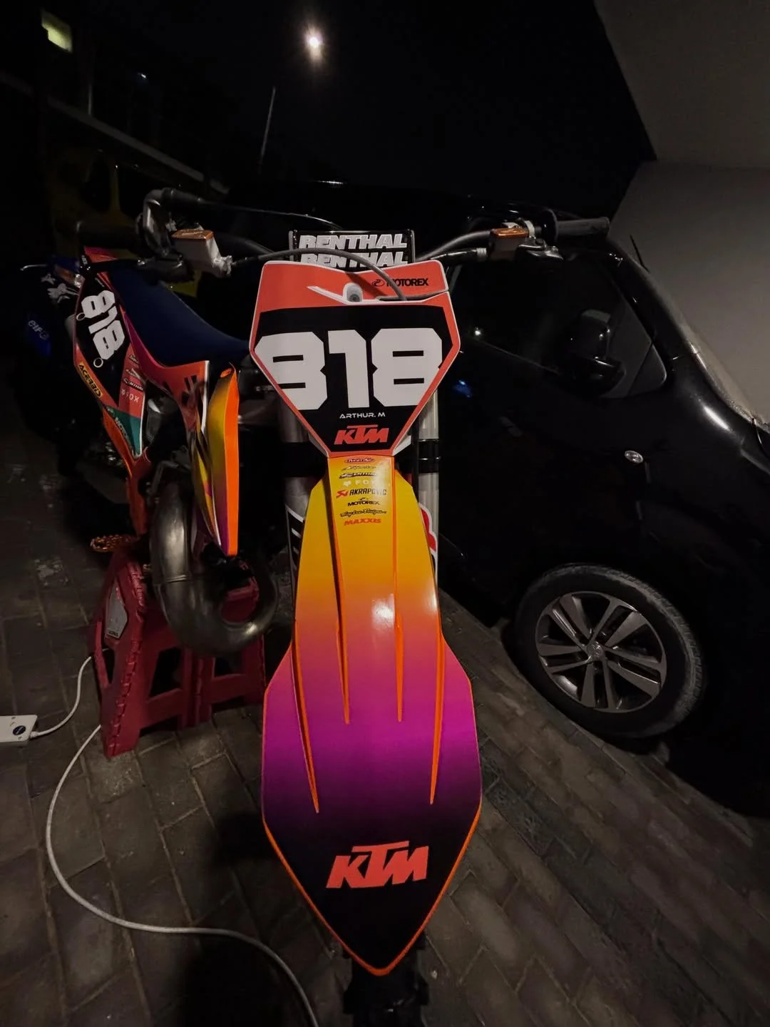 KTM dirt bike with front number plate 318, colorful front fender, and sponsor stickers, parked next to a black vehicle.