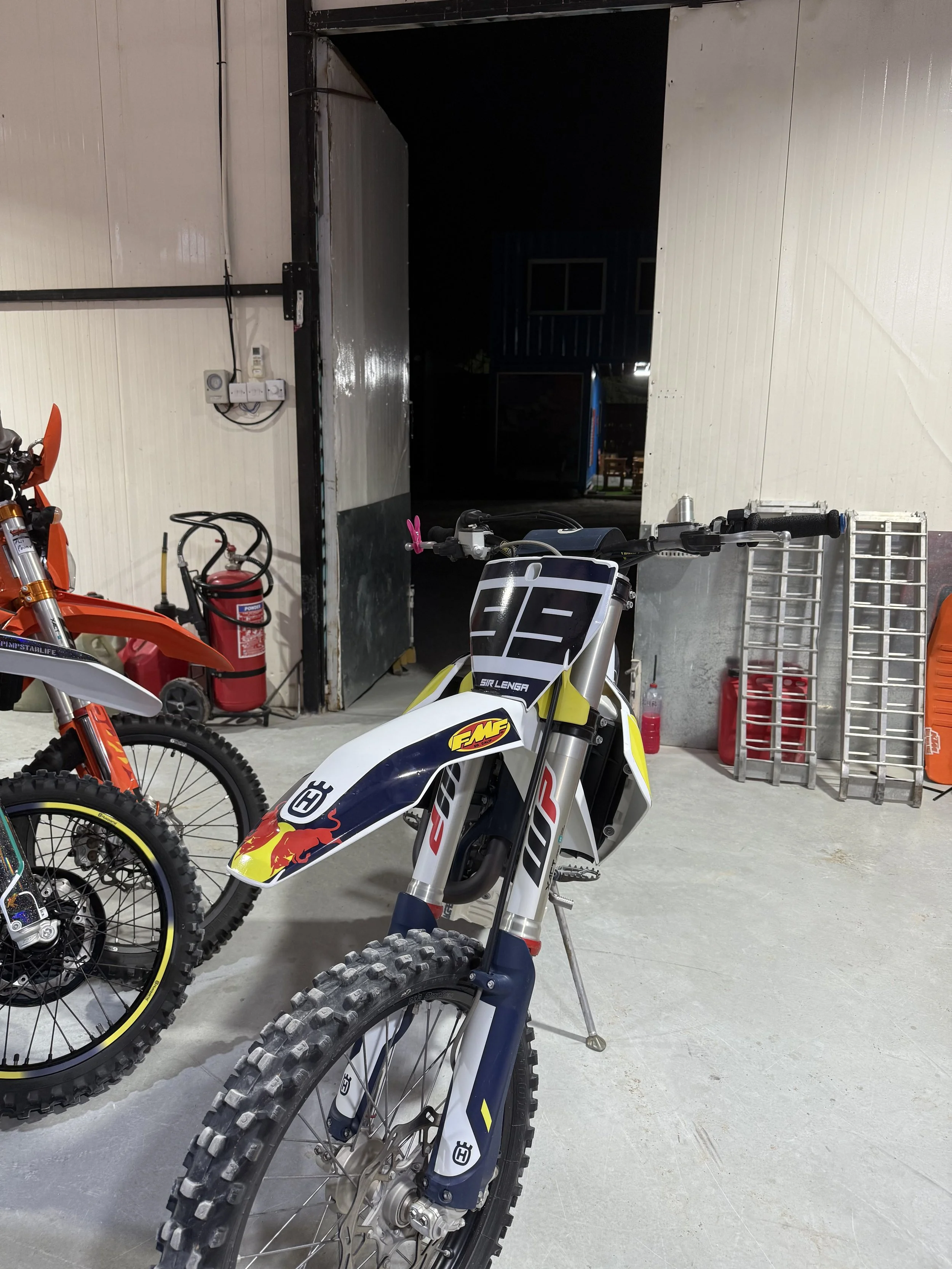 Indoor garage with two motocross bikes, a fire extinguisher, and a ladder positioned near a building wall. The garage has white paneled walls, a concrete floor, and an open roller door leading outside, revealing a dark night and a vending machine.