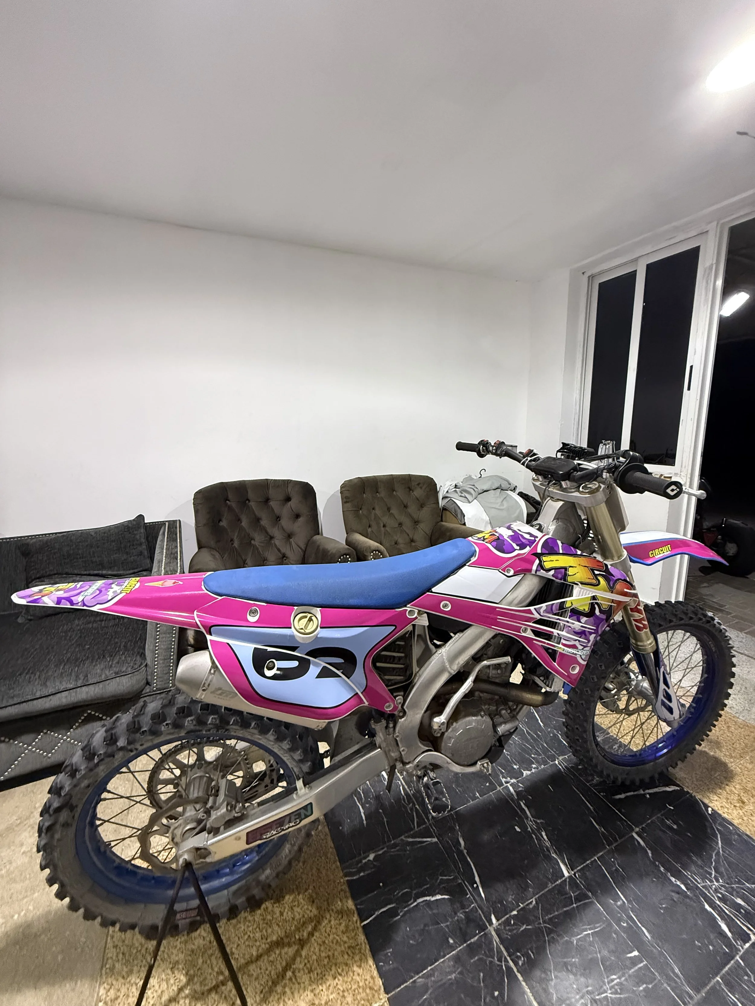 A motocross dirt bike with pink, purple, and white graphics is placed indoors, partially blocking a black sofa. Behind it, there are two brown tufted chairs and a window with white framing. The room has a black and white marble-patterned floor.