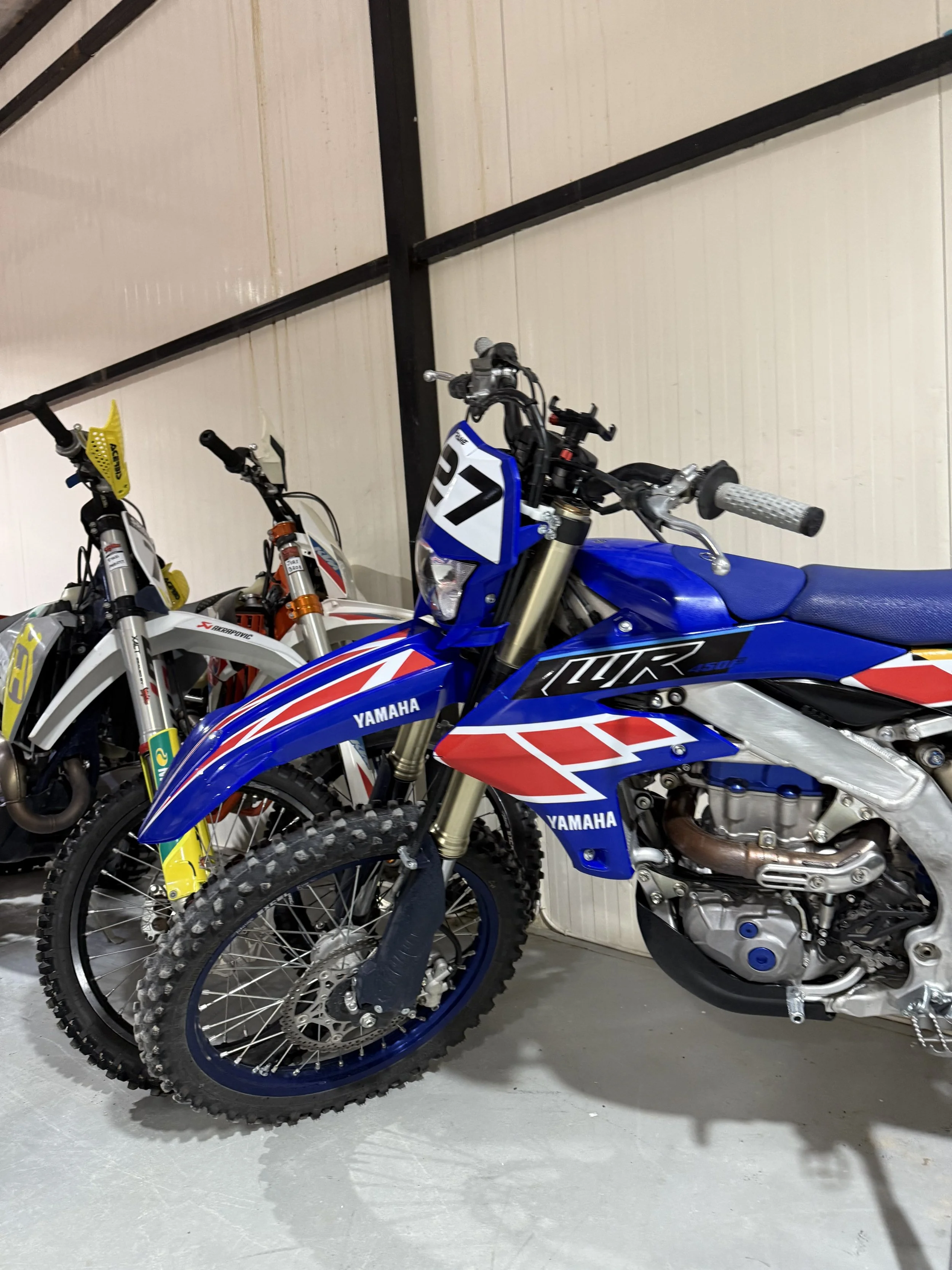Multiple dirt bikes are lined up indoors, with the front bike being a Yamaha model in blue with racing numbers. The bikes have knobby tires, and some have yellow or orange accent parts. The background features a wall with black metal beams.
