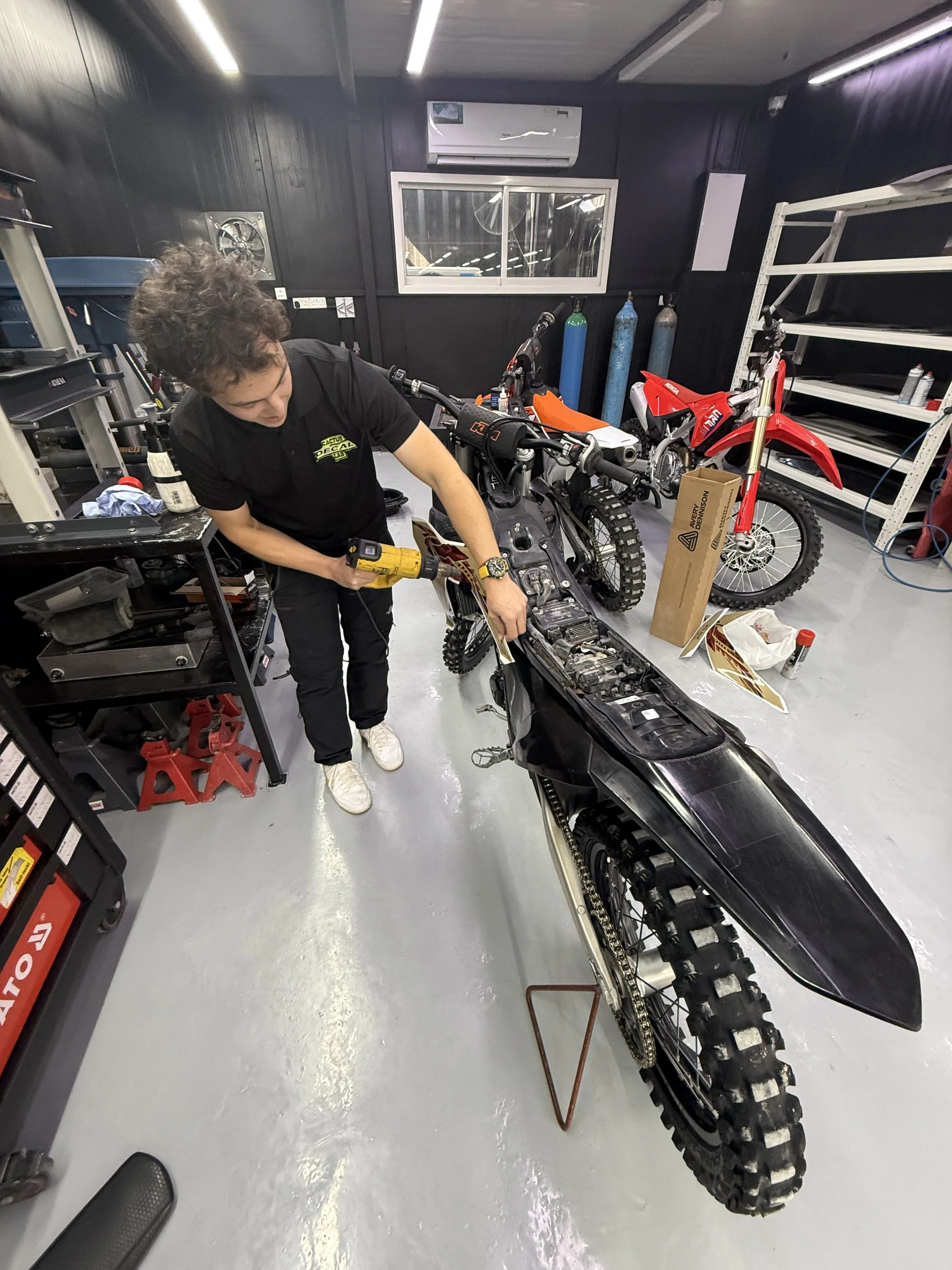 A person working on a dirt bike in a workshop. The workshop has black walls, a window, and various tools and equipment. There are two gas cylinders in the background, a box, and shelves on the right side.