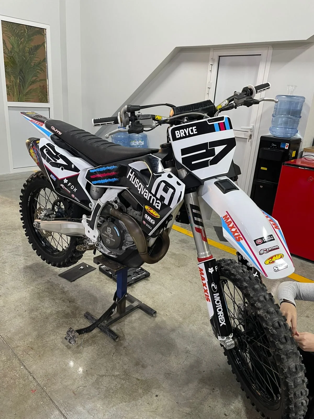 Racing dirt bike with sponsor decals, labeled 'Bryce', inside a workshop with water cooler and red equipment in background.