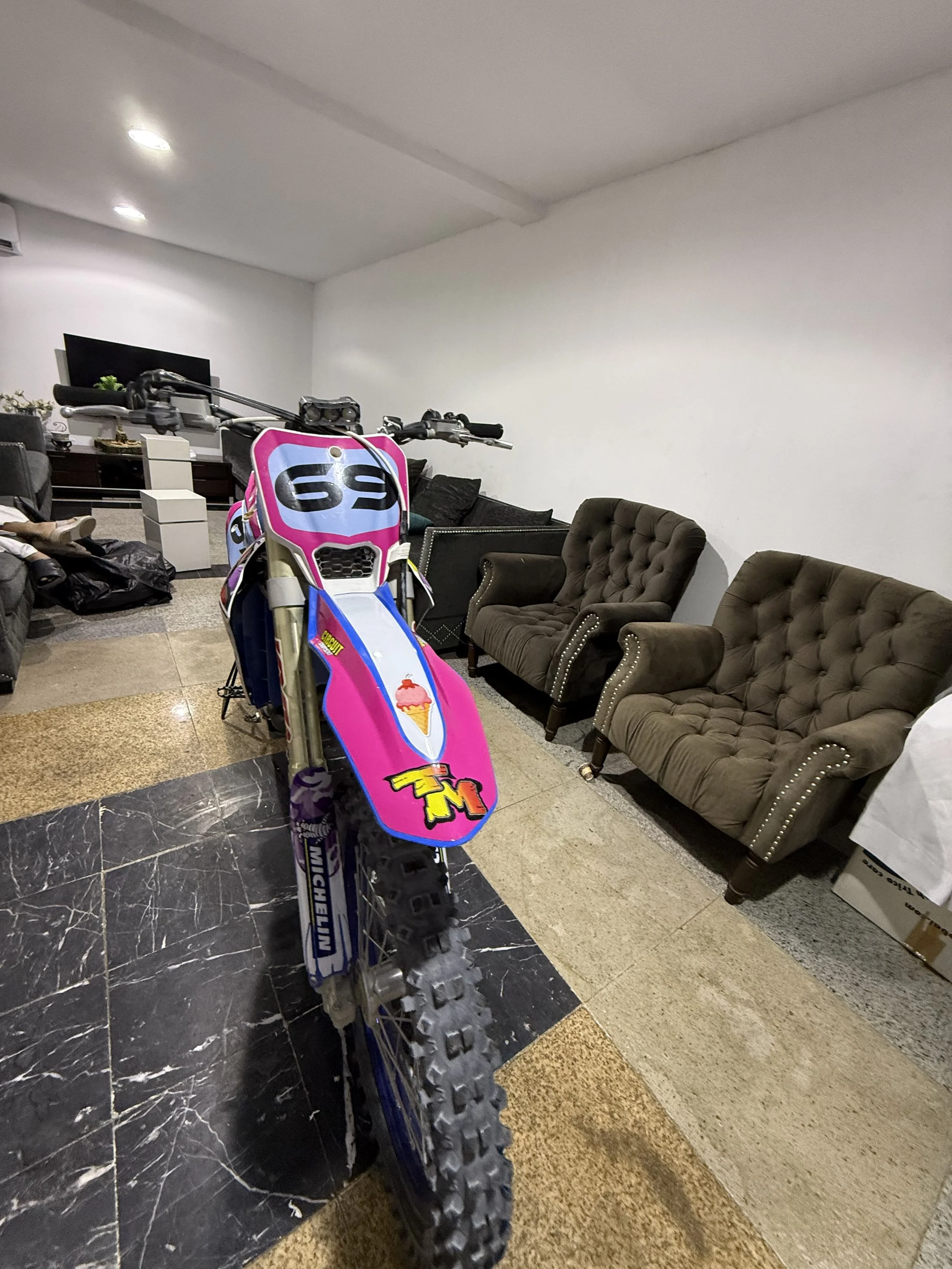 Indoor room with leather cushioned chairs, a sofa, a TV on a white stand, and a pink and purple dirt bike with racing decals in the foreground.