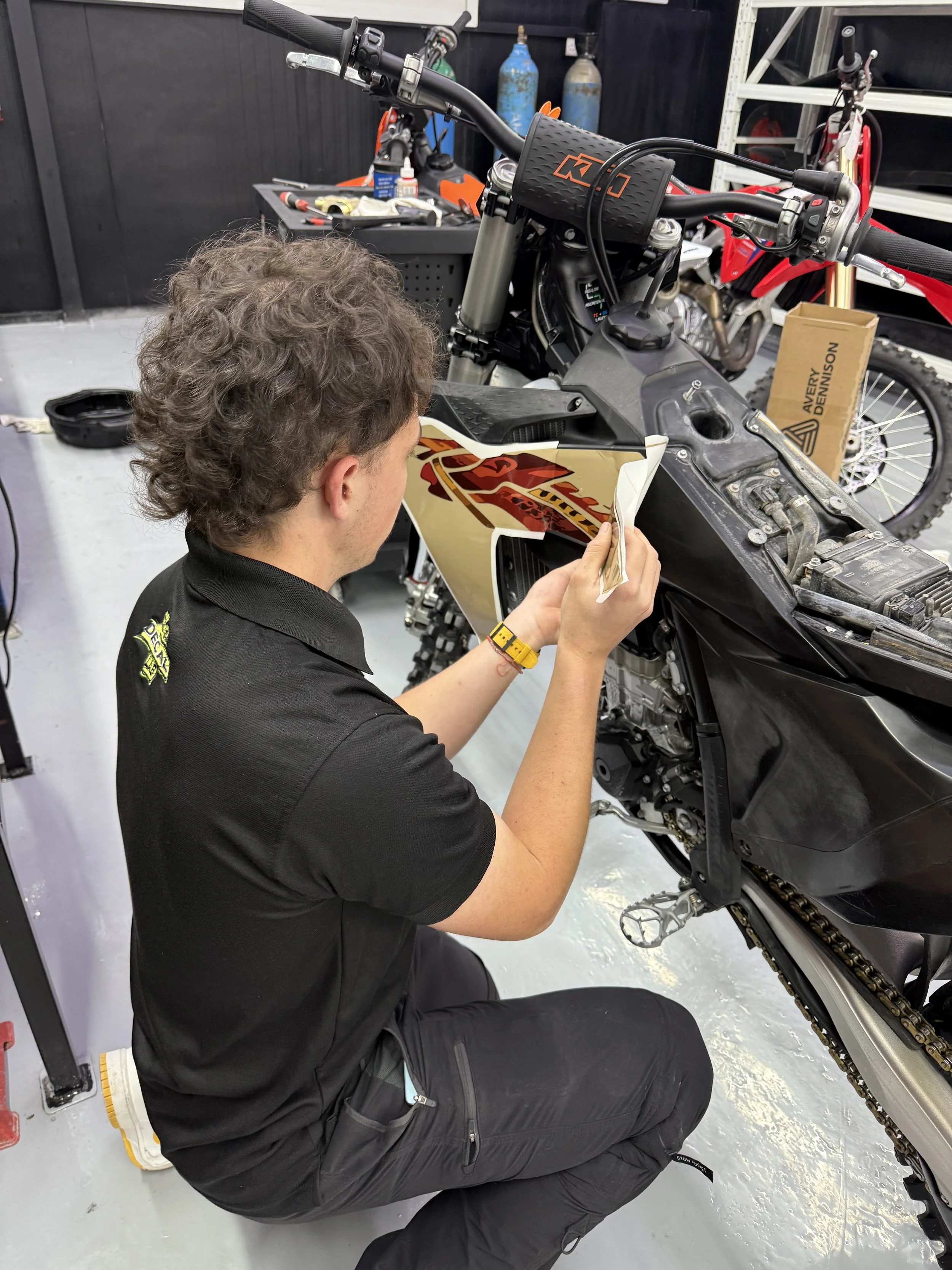 A person with curly hair, wearing a black shirt with a yellow logo on the sleeve, is kneeling and applying decals to a black and orange dirt bike in a garage or workshop.
