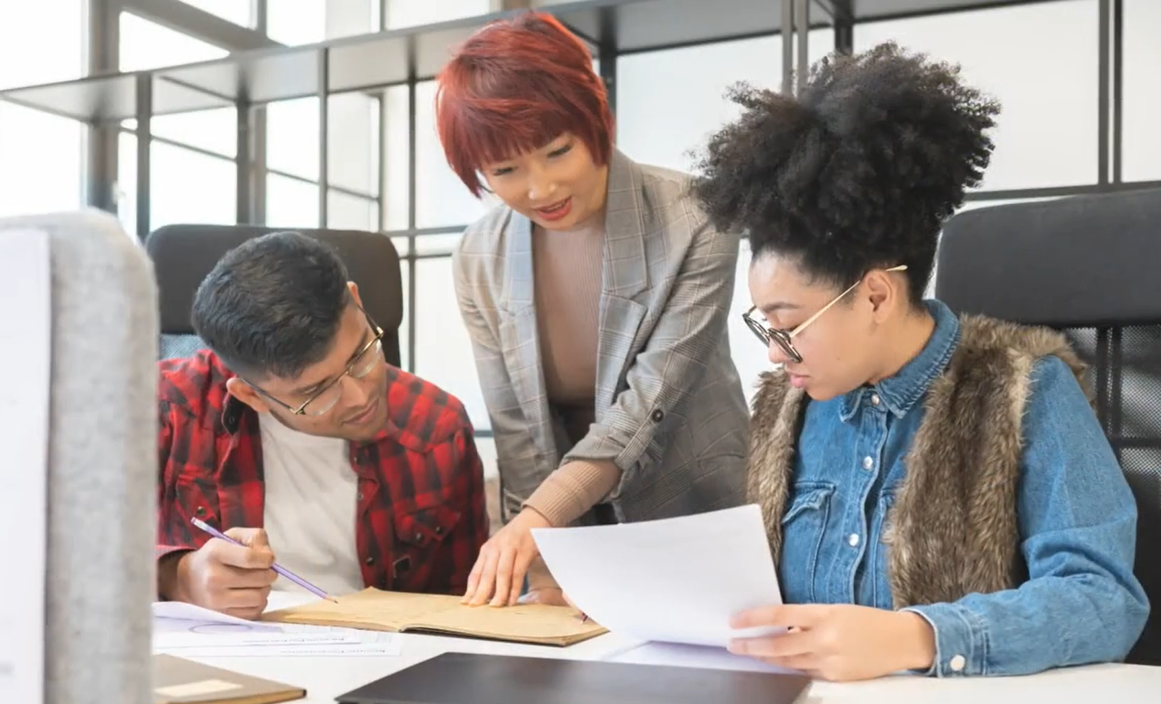 A woman with short red hair in a gray blazer is helping two young women with documents at a desk in an office setting.