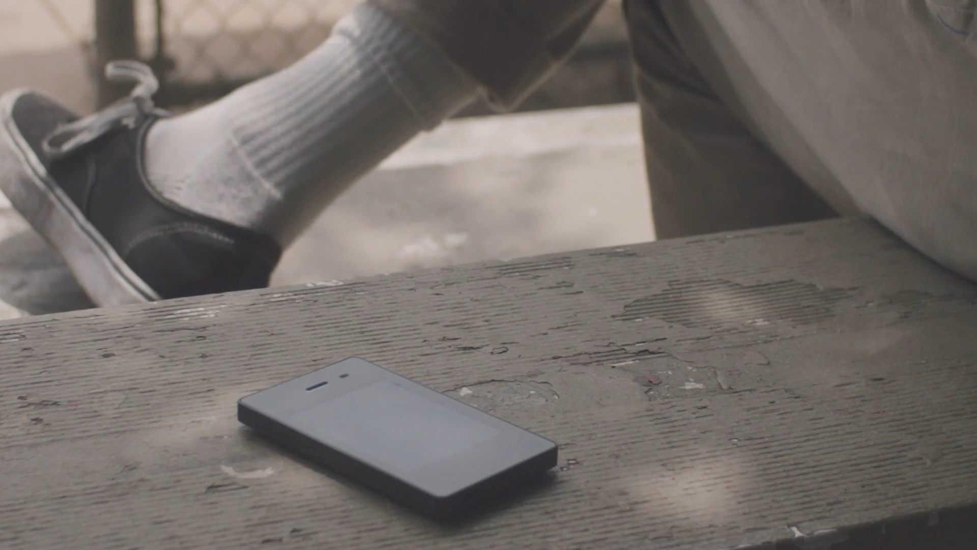 Close-up of a wooden table with a smartphone and a person's legs visible in the background, wearing gray socks and black shoes.