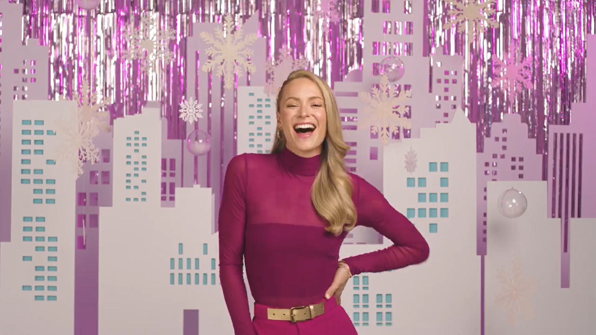 A woman with long blonde hair, smiling and wearing a fuchsia long-sleeve turtleneck and matching pants with a beige belt, standing in front of a festive holiday backdrop with pink and purple cityscape, snowflakes, and hanging ornaments.