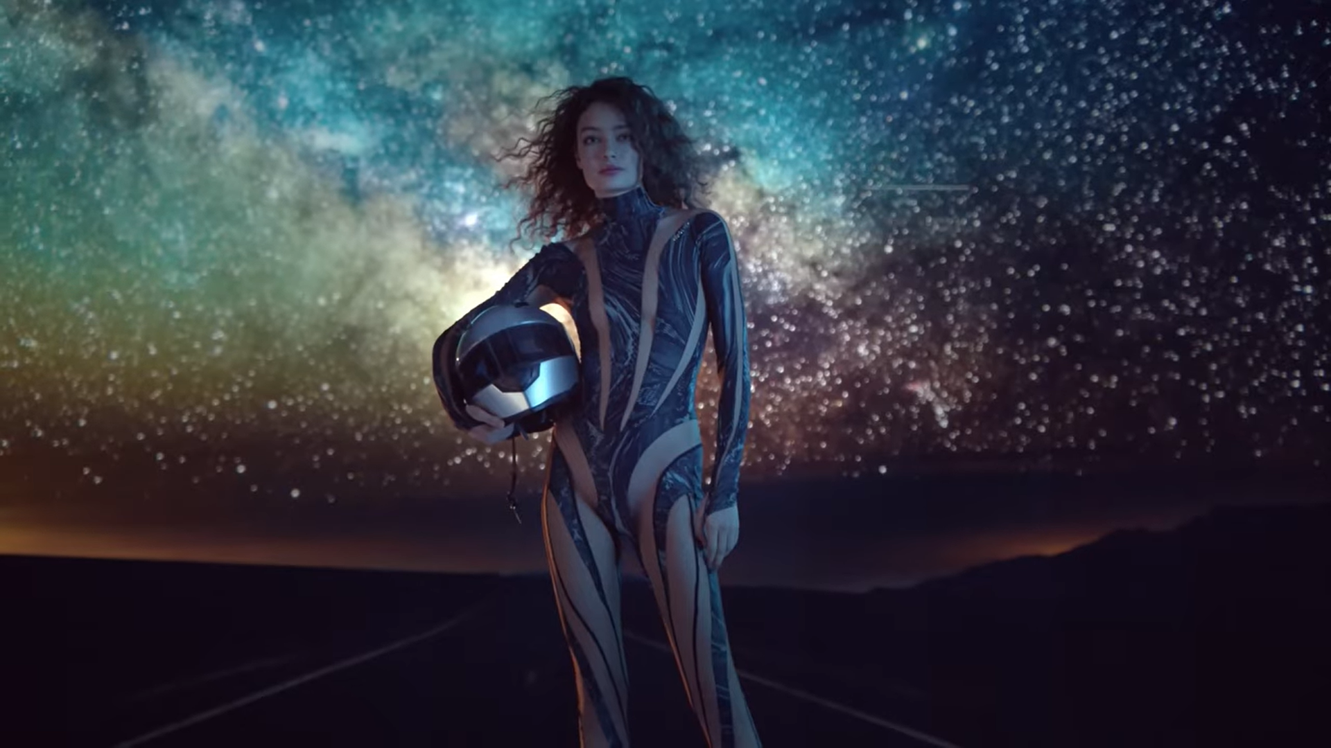 A woman in a futuristic, patterned bodysuit stands under a star-filled night sky, holding a helmet, with a mountain landscape in the distance.