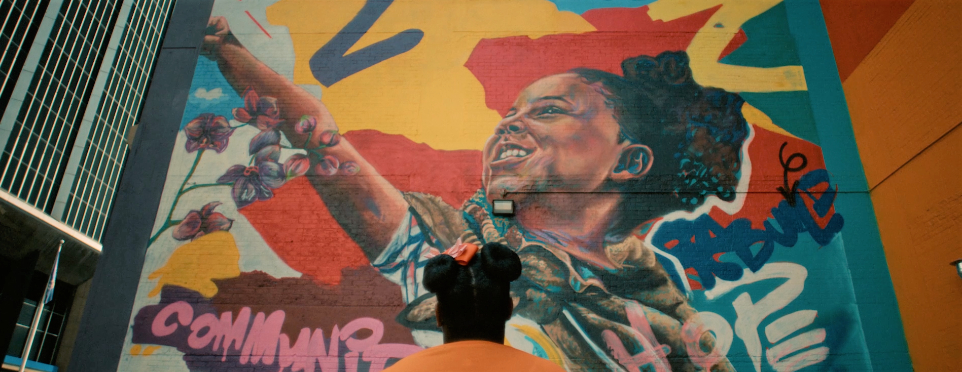 Back of a person with a pink bow in their hair, standing in front of a colorful mural of a woman with braids, flowers, and a joyful expression, painted on a brick wall.