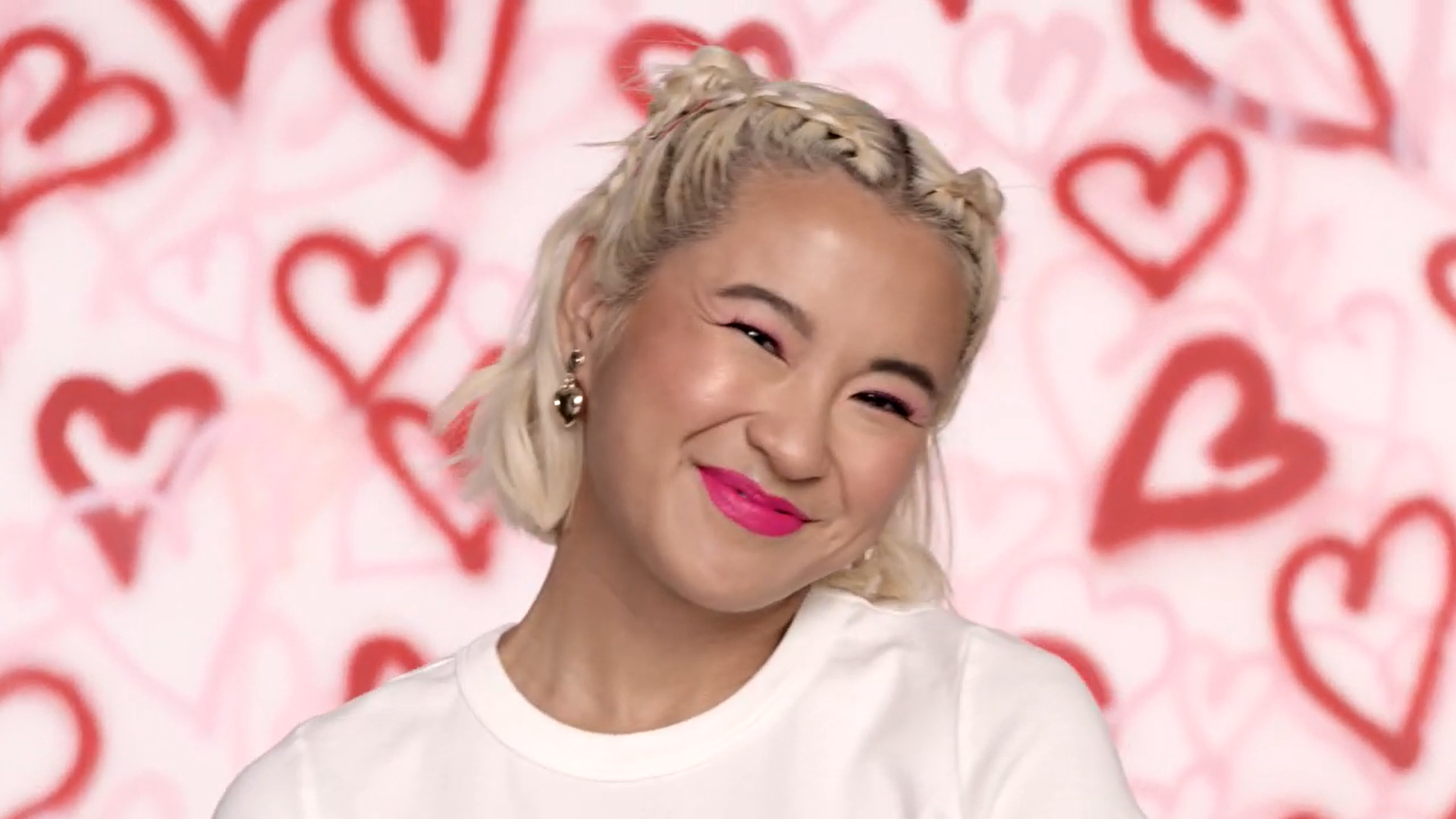 A woman with blonde hair styled in braids and buns, smiling with pink lipstick, wearing earrings and a white shirt, standing in front of a pink background with red heart patterns.