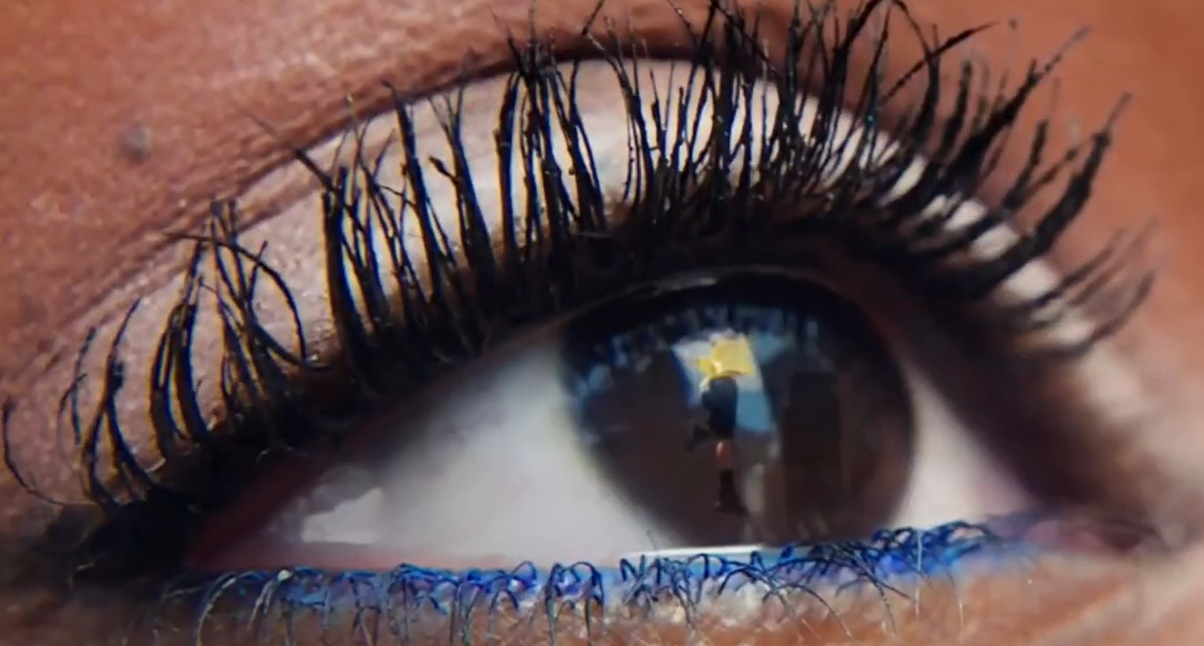 A close-up of a human eye with long, dark eyelashes, brown skin, and a reflection in the eye showing the person taking the photo with a yellow umbrella.