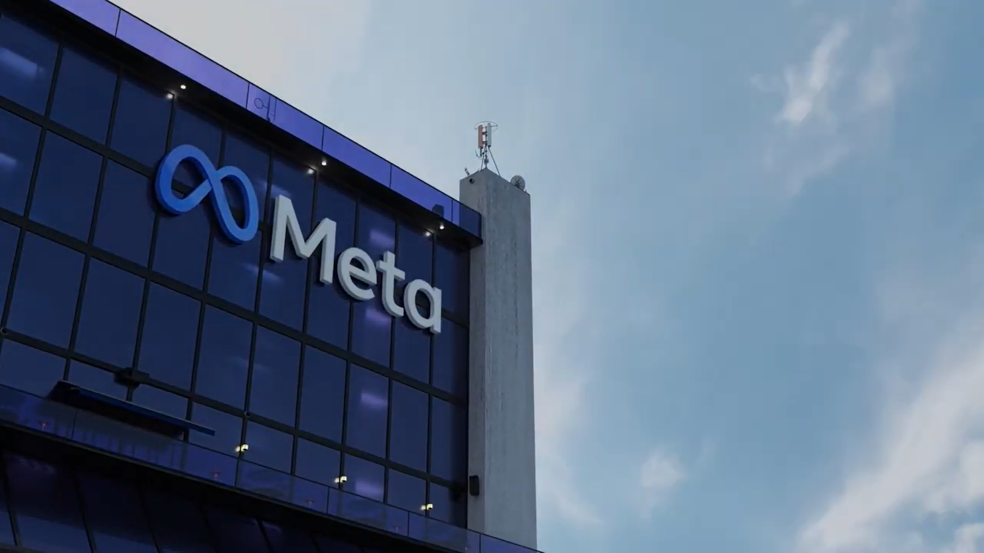 Exterior of a building with a glass facade displaying the Meta logo and name, under a partly cloudy sky, with a communication antenna on the rooftop.